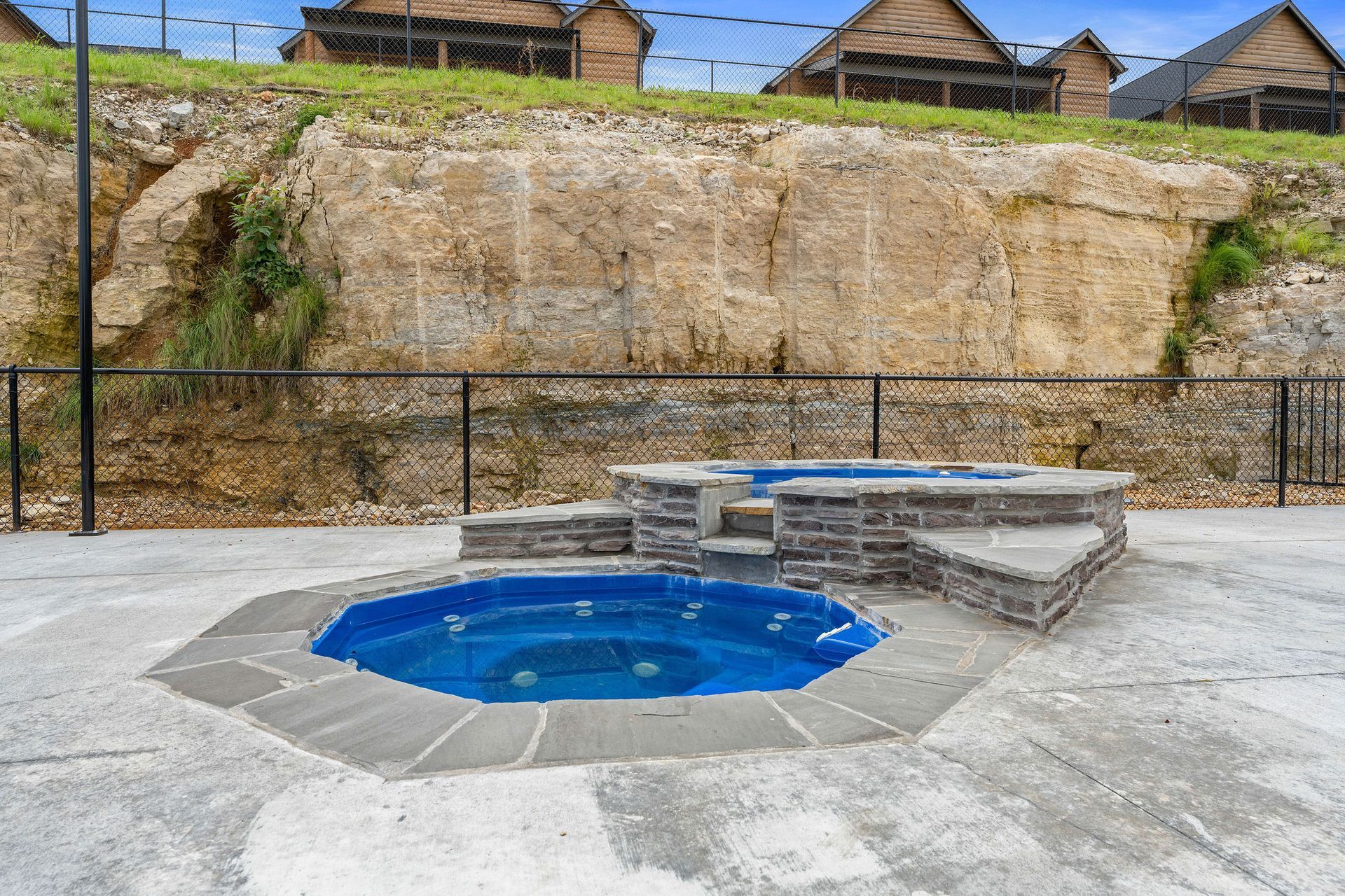 Small blue hot tub in a concrete courtyard beside stone benches and a rocky hillside backdrop