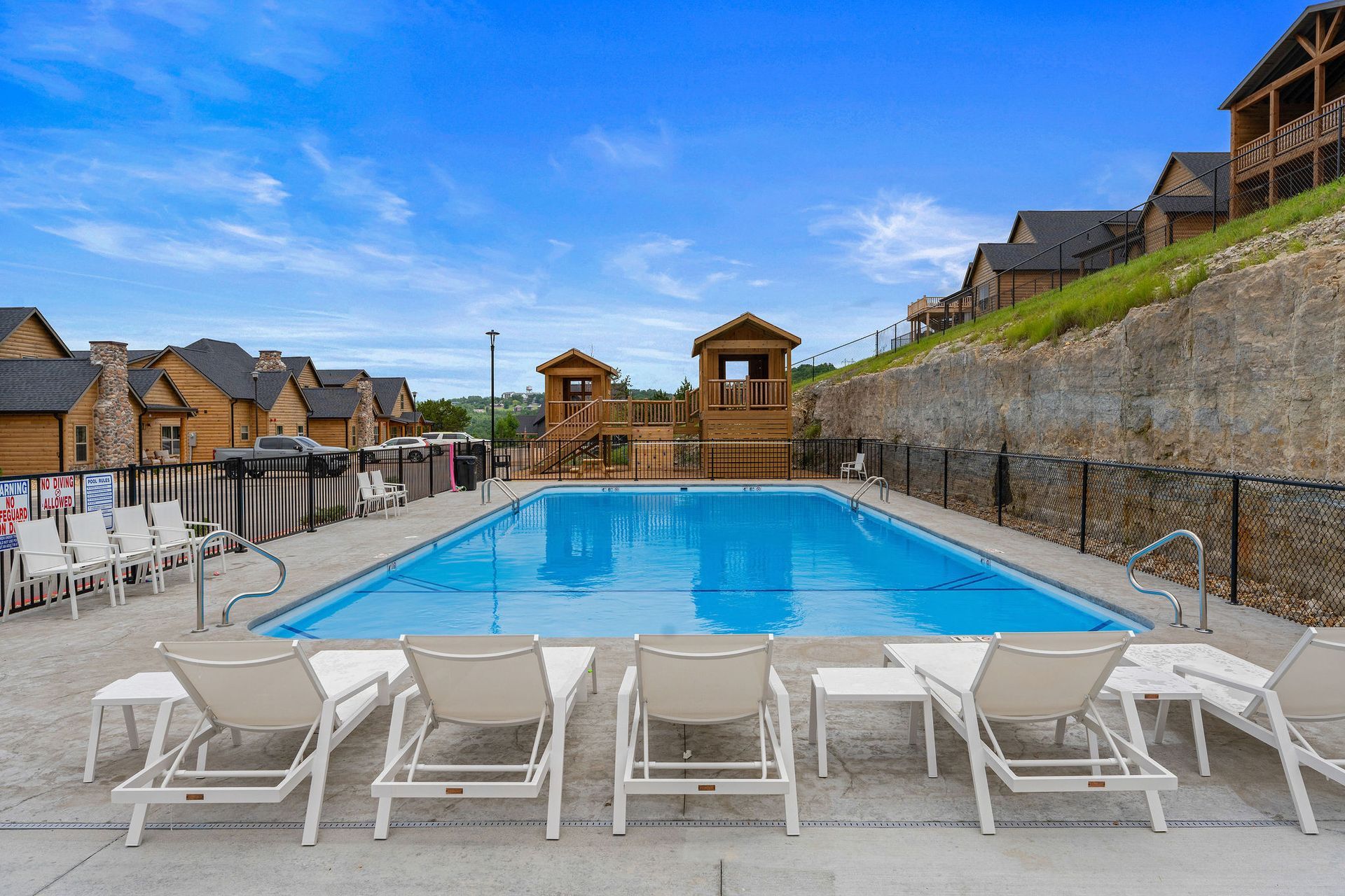 Outdoor swimming pool with white lounge chairs, stone walls, and resort buildings under a blue sky