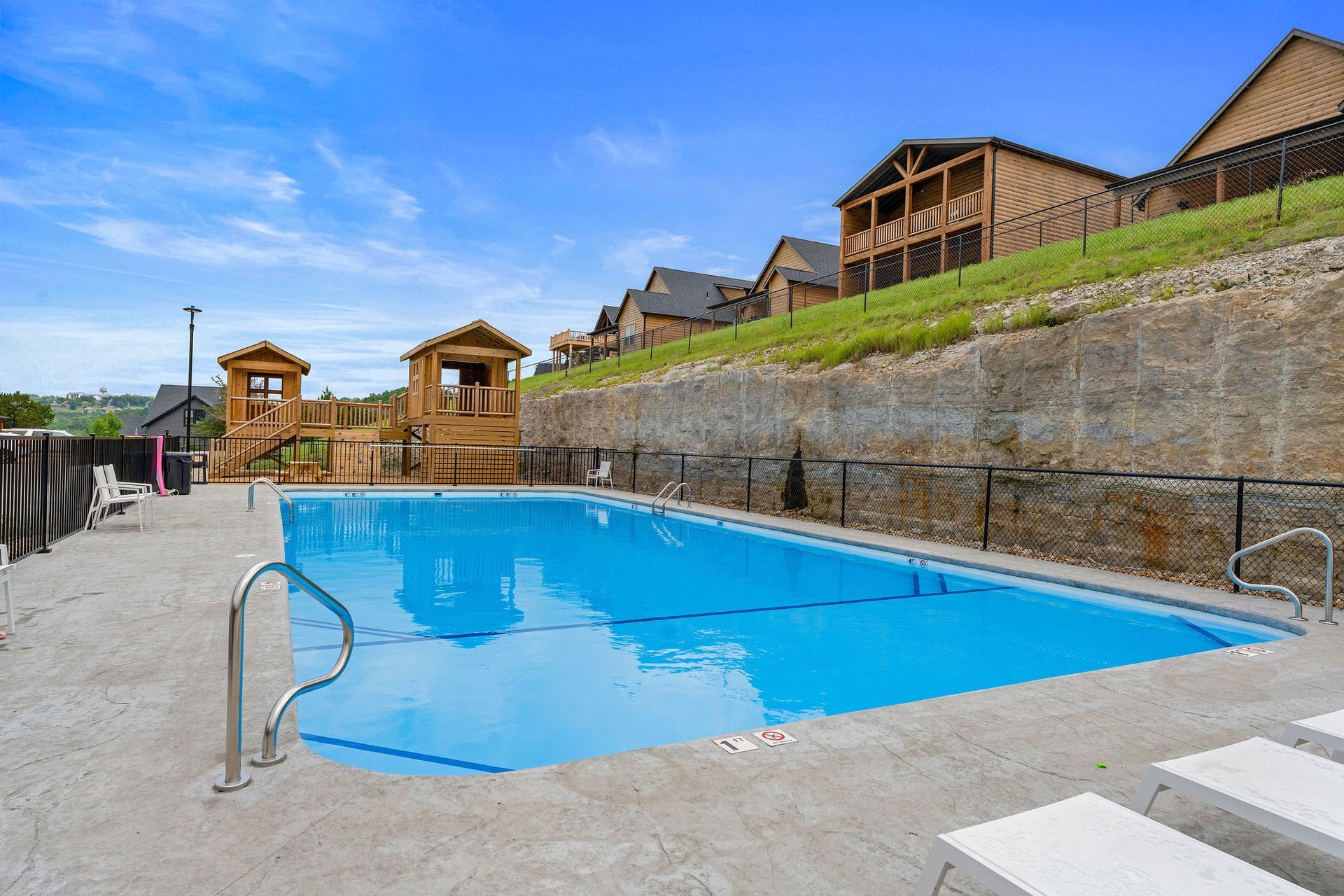 Outdoor swimming pool beside a stone wall and hillside cabins under a blue sky