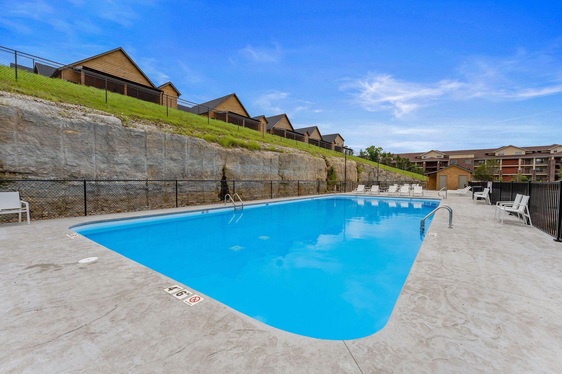 Outdoor swimming pool beside a stone retaining wall, with blue water and houses on a grassy hillside.