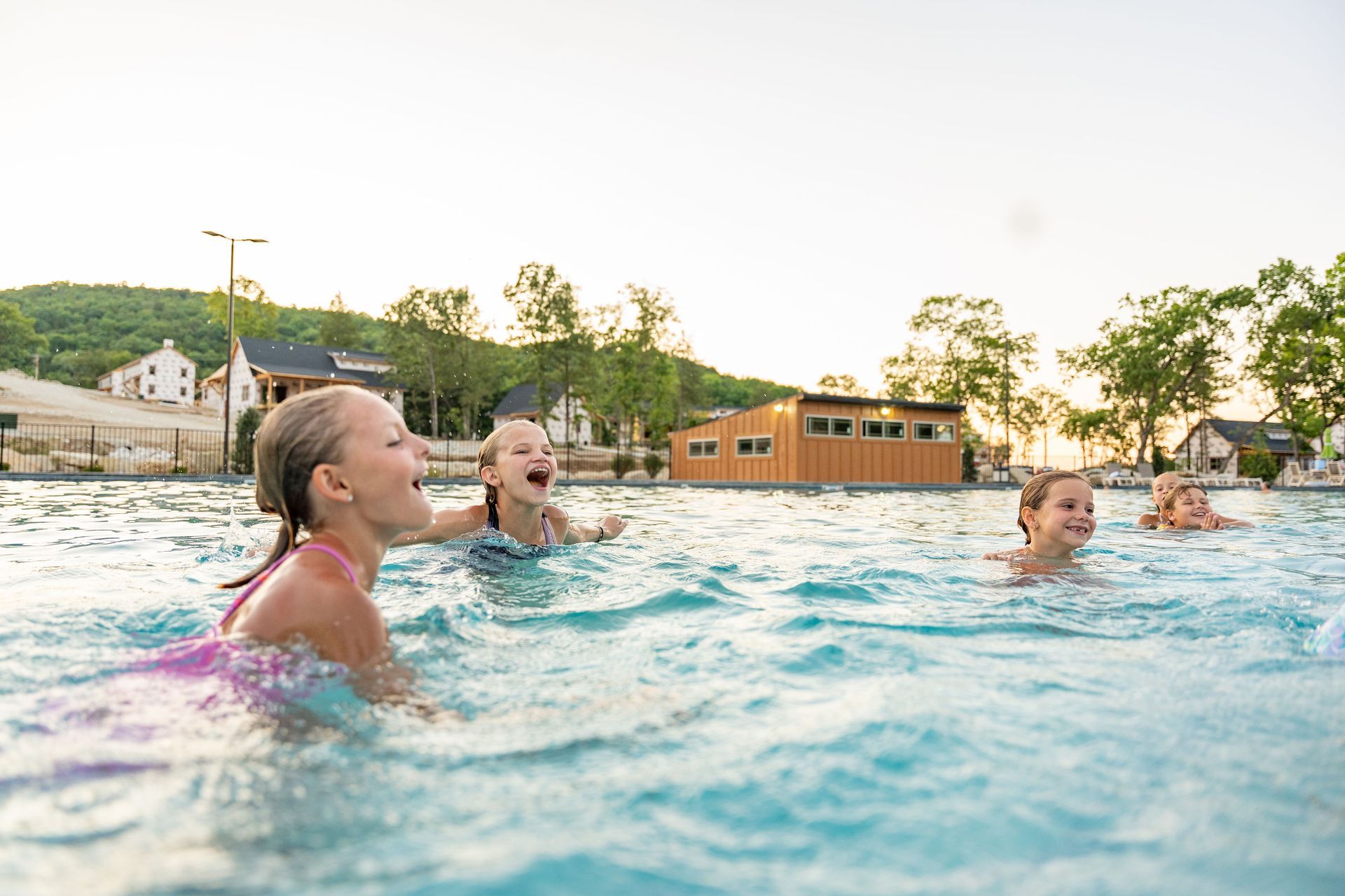 People swimming in an outdoor pool at sunset, with buildings and trees in the background
