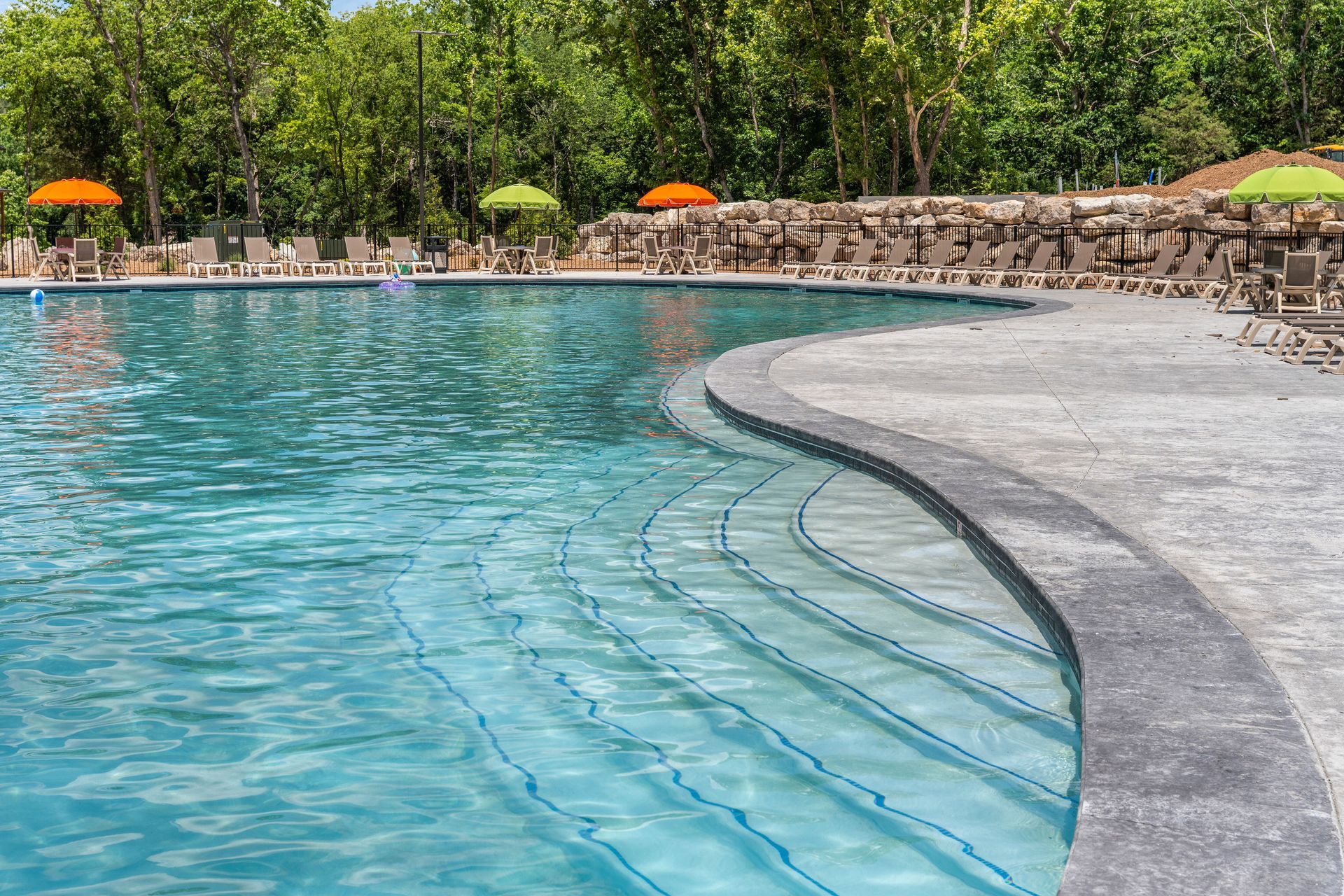 Curved outdoor swimming pool with turquoise water, stone edge, and lounge area under orange umbrellas in a leafy setting