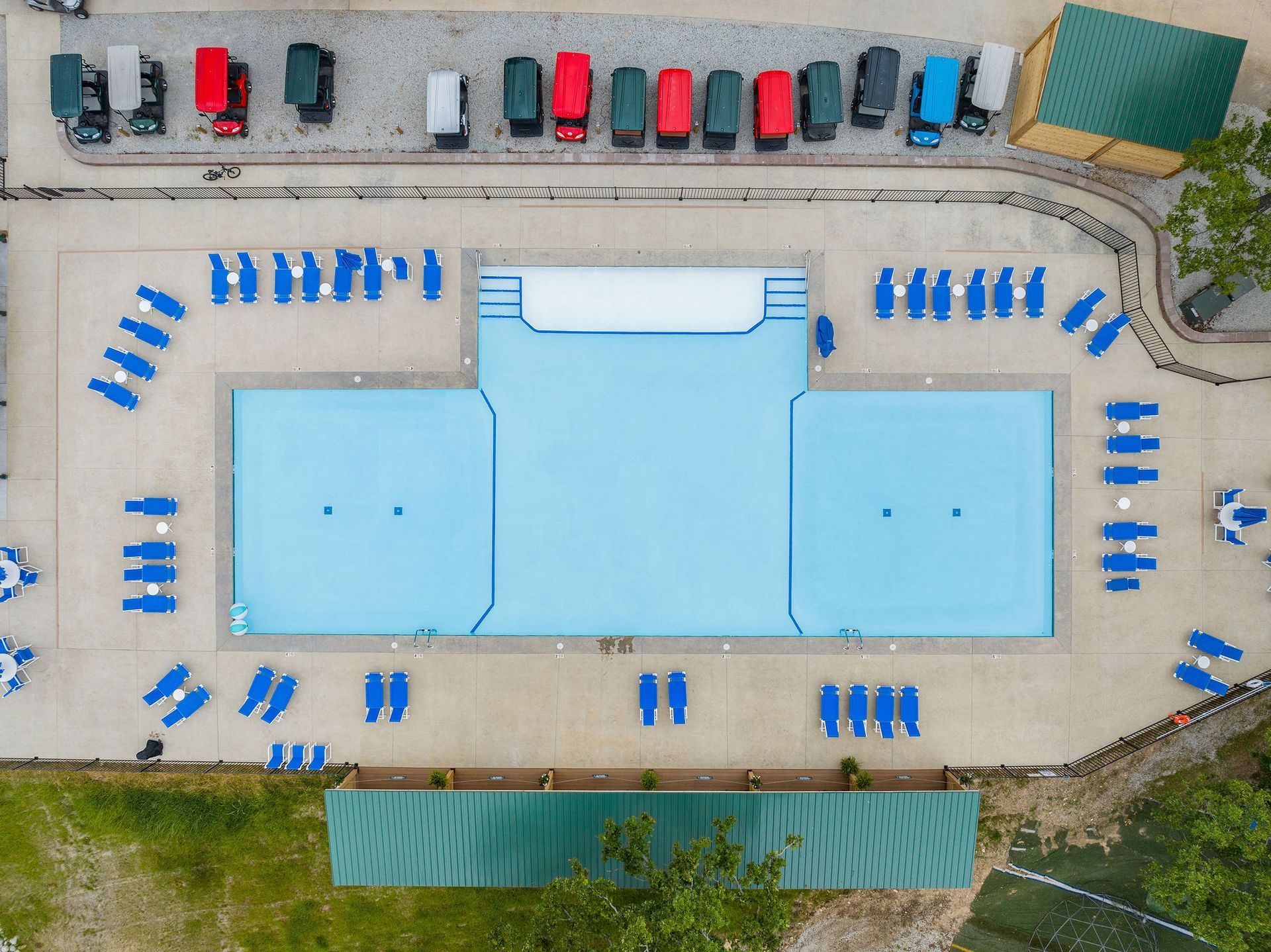 Aerial view of an outdoor swimming pool with blue lounge chairs and parked cars nearby.