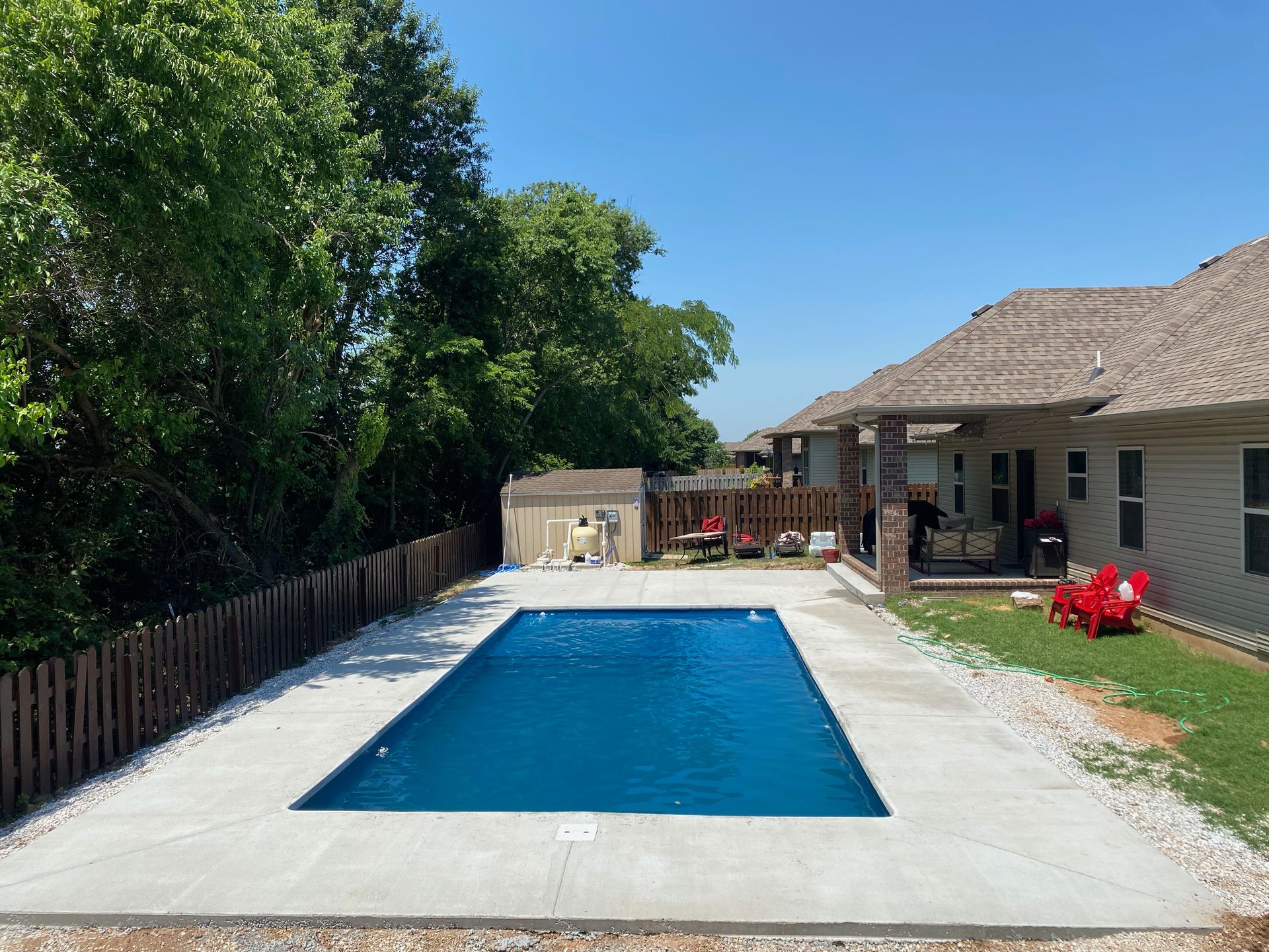 Backyard swimming pool beside a house and patio, with trees and a clear blue sky.