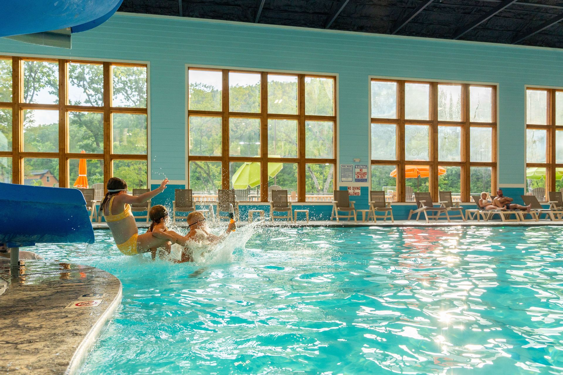Indoor pool with people splashing near large windows and lounge chairs