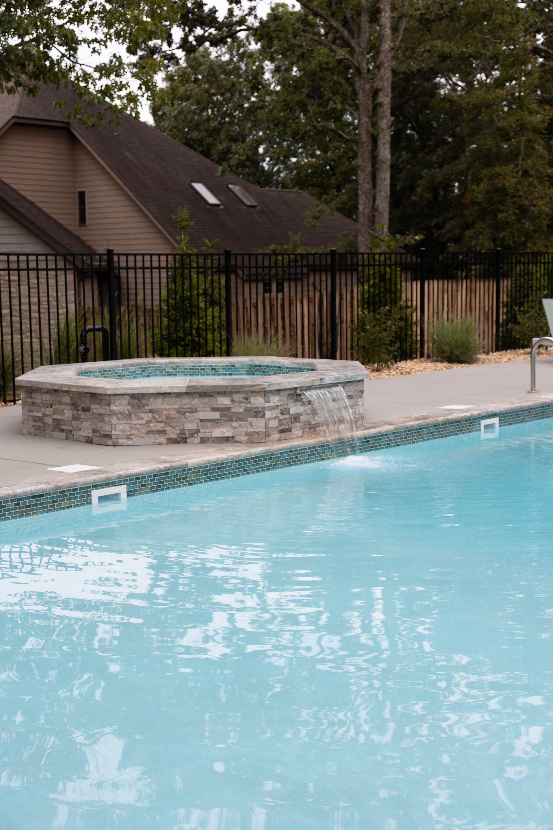 Outdoor swimming pool with a stone hot tub, black fence, and trees nearby