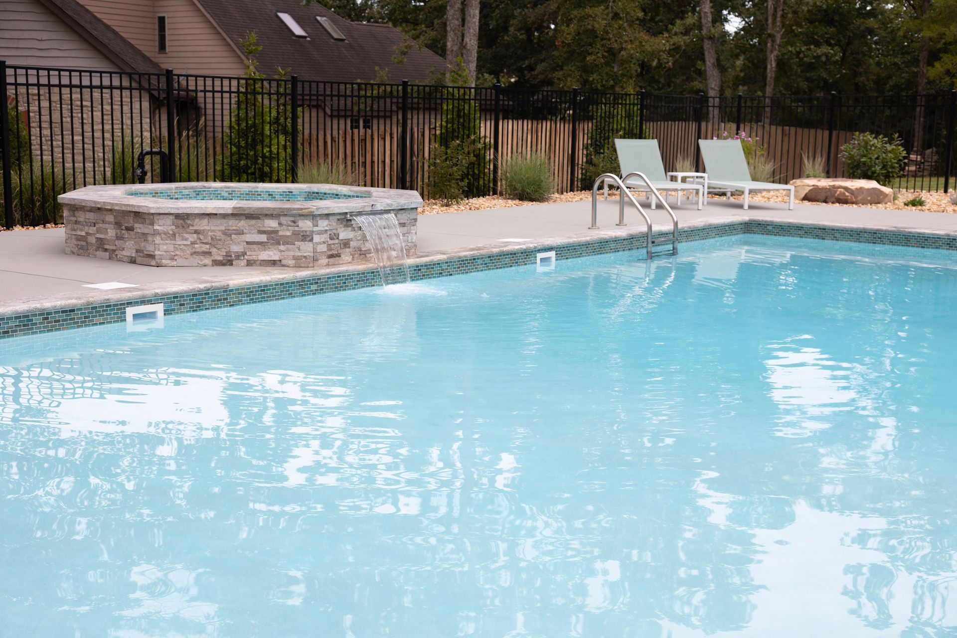 Outdoor swimming pool with a stone hot tub, lounge chairs, and a fence in the background