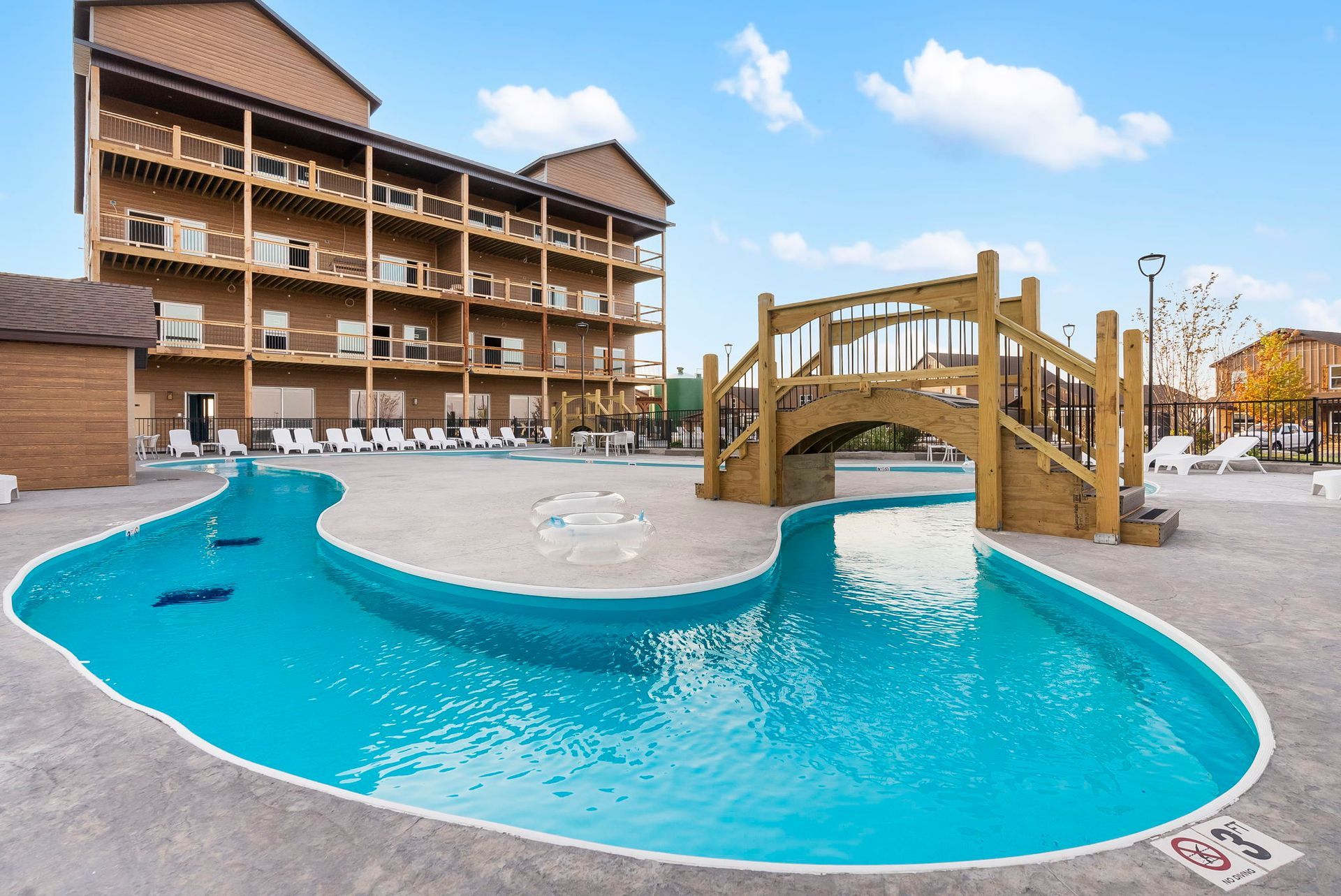 Outdoor resort pool with a wooden bridge and hotel balconies under a blue sky