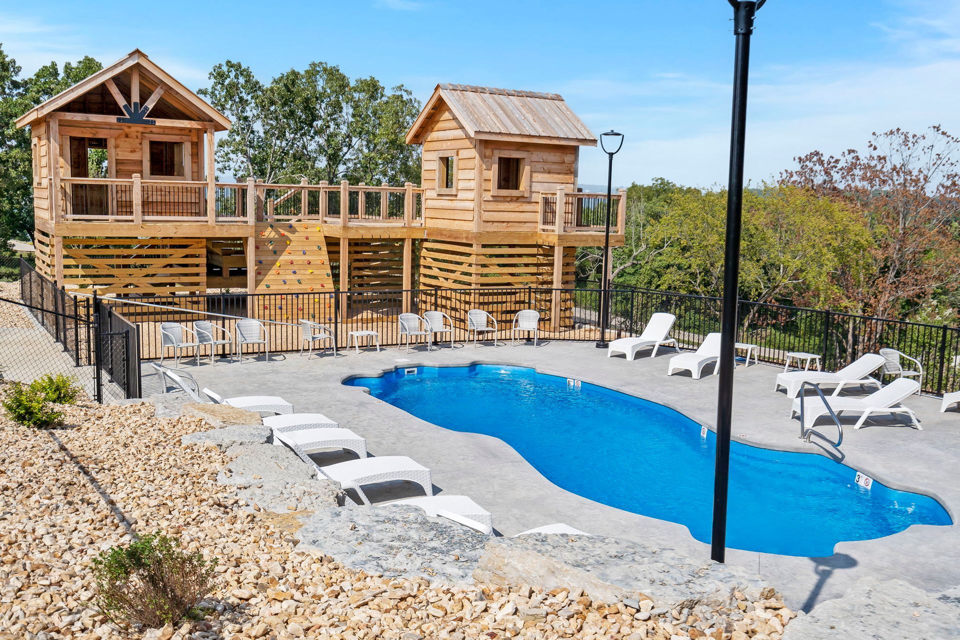 Outdoor pool area with two wooden cabins, lounge chairs, and blue water surrounded by gravel and trees