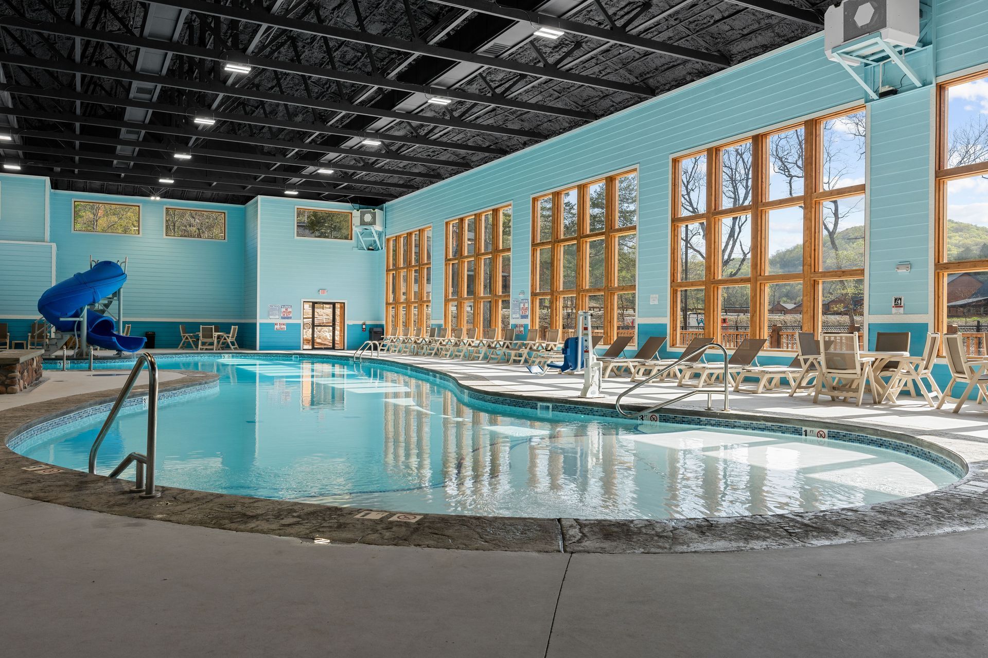 Indoor swimming pool with blue water, lounge chairs, large windows, and a water slide