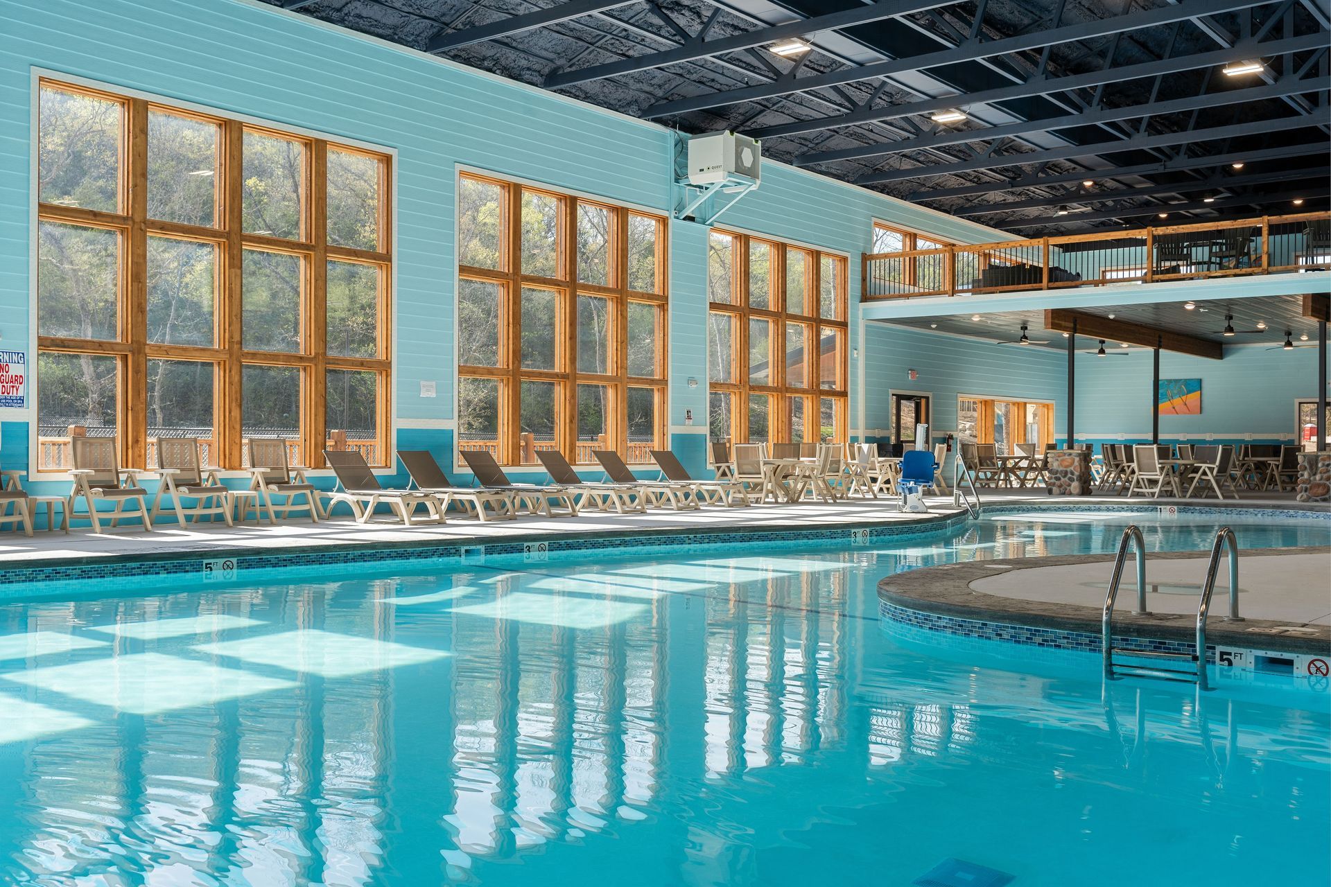 Indoor swimming pool with turquoise water, lounge chairs, and tall windows reflecting sunlight