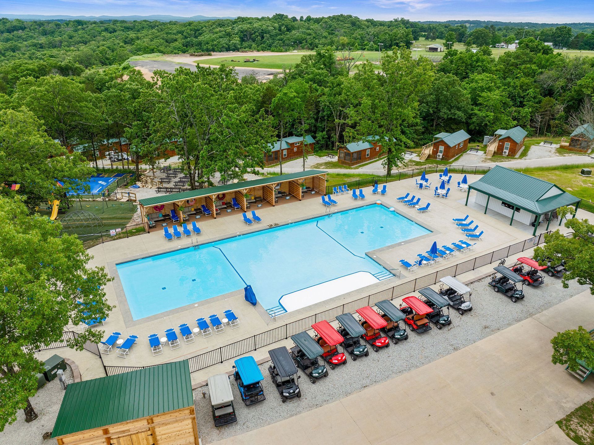 Aerial view of an outdoor pool complex with blue pools, lounge chairs, and red canopy carts amid trees.
