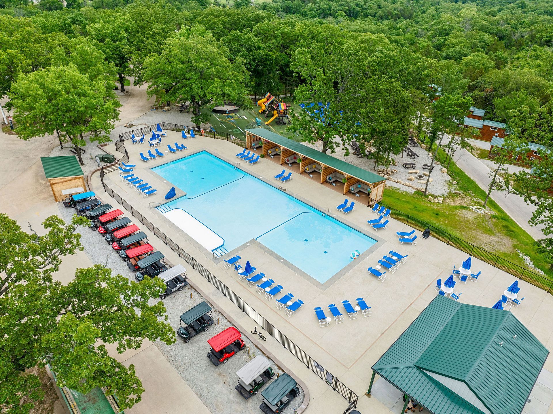 Aerial view of an outdoor pool with blue lounge chairs, trees, parked cars, and a pavilion nearby