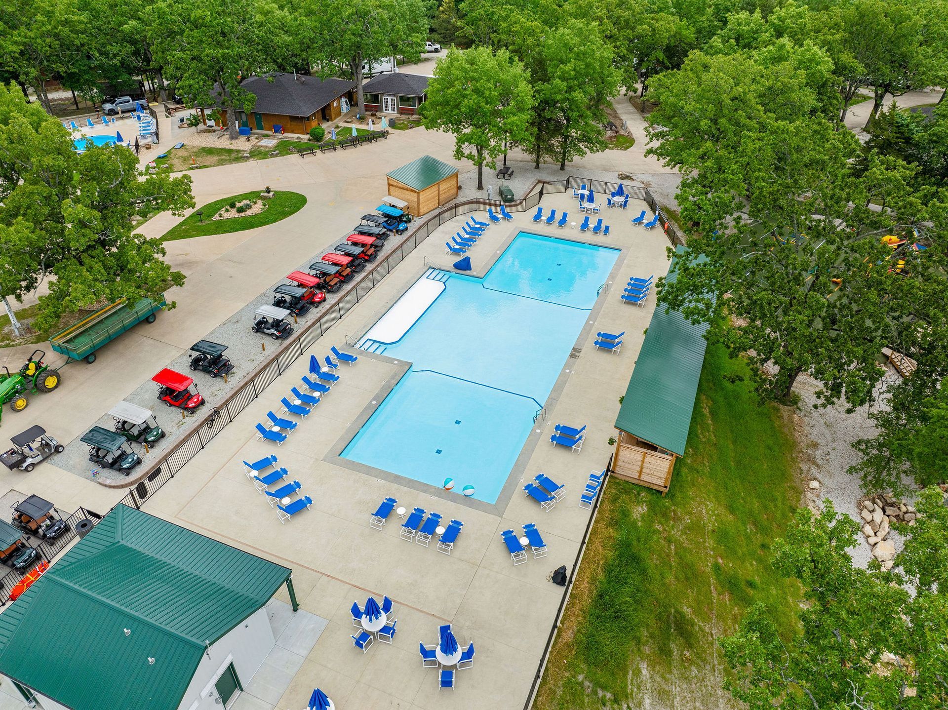 Aerial view of an outdoor swimming pool with blue lounge chairs, parked cars, and trees surrounding it
