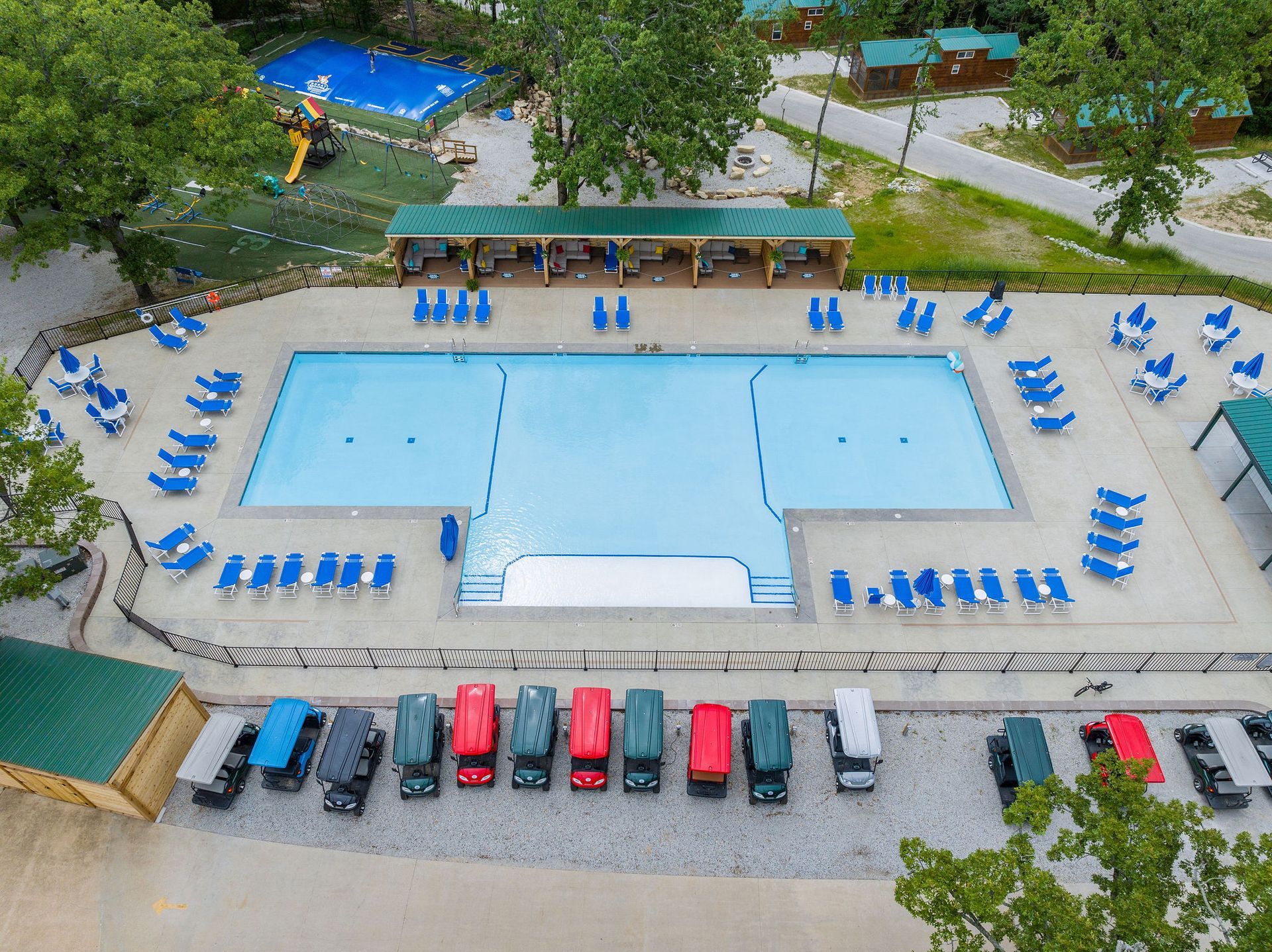 Aerial view of a resort swimming pool with blue lounge chairs, shaded cabanas, and parked cars nearby