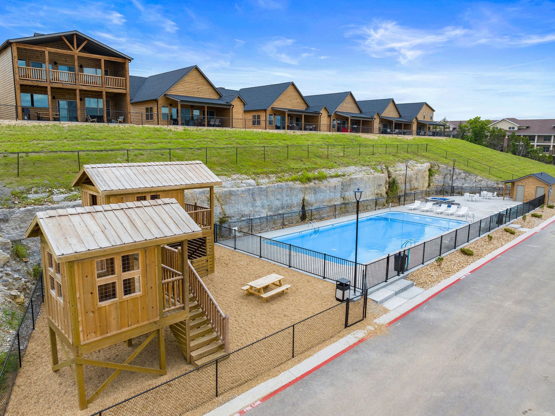 Resort-style outdoor pool beside wooden cabins on a grassy hillside, with a road in the foreground
