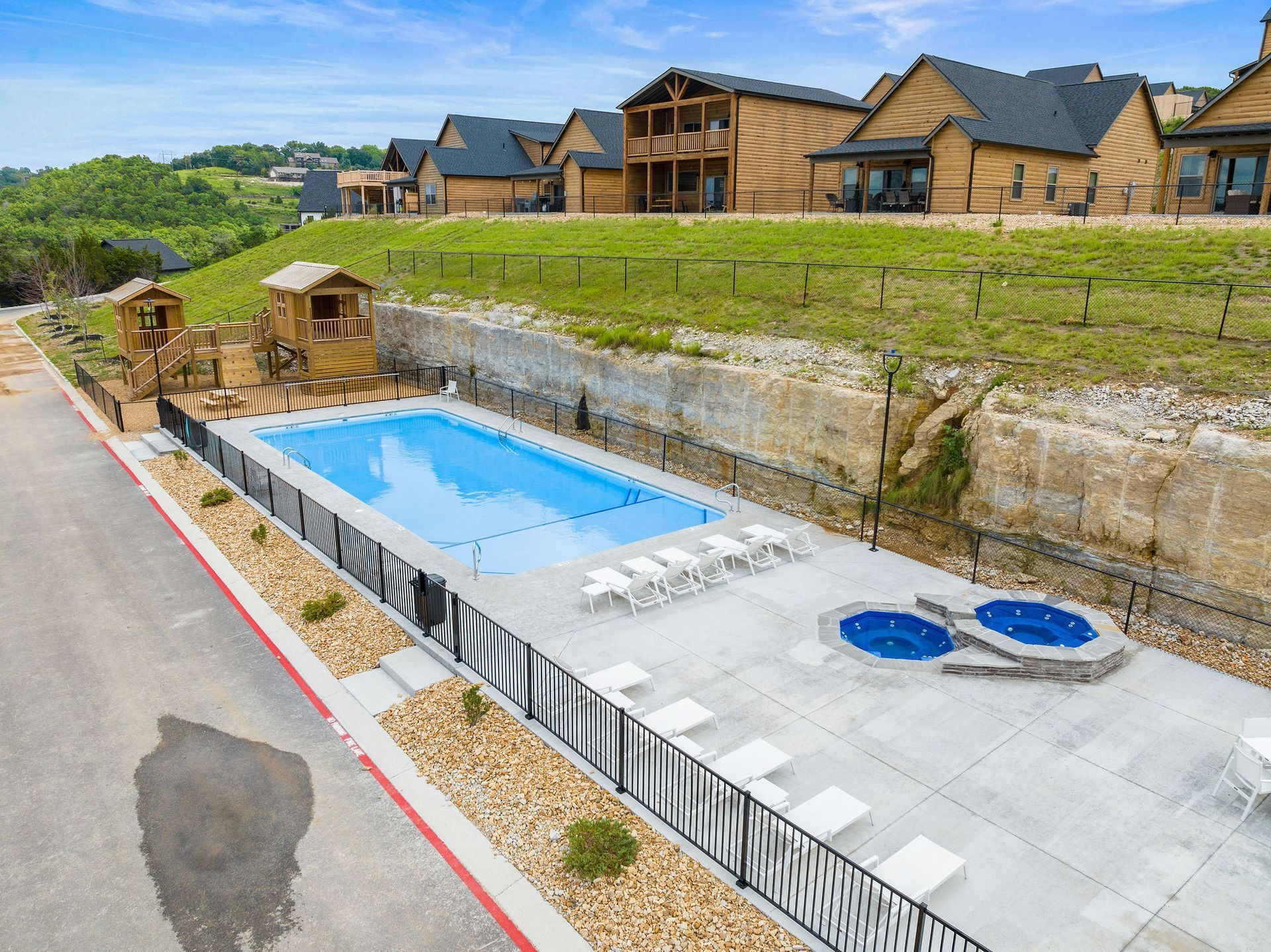 Outdoor pool area beside modern wooden houses, with a blue pool and two round hot tubs on a concrete deck.