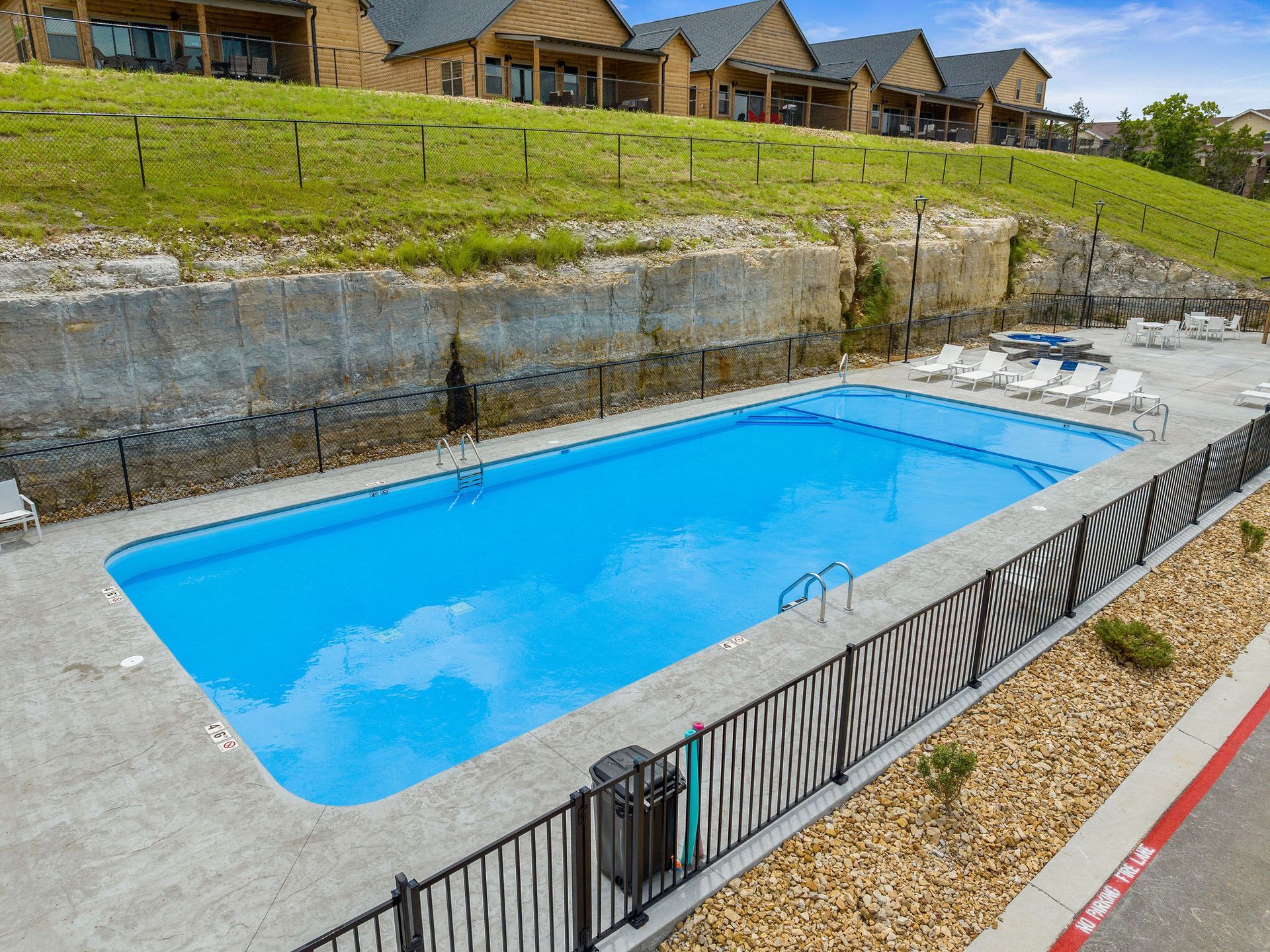 Outdoor swimming pool beside a sloped lawn and row of houses, with a fence and cloudy water.