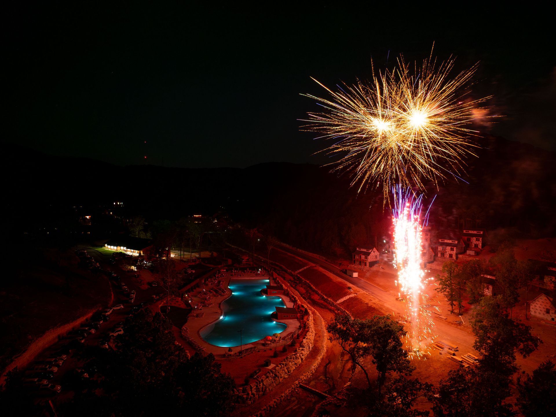 Nighttime fireworks above a glowing blue pool and lit paths in a dark landscape