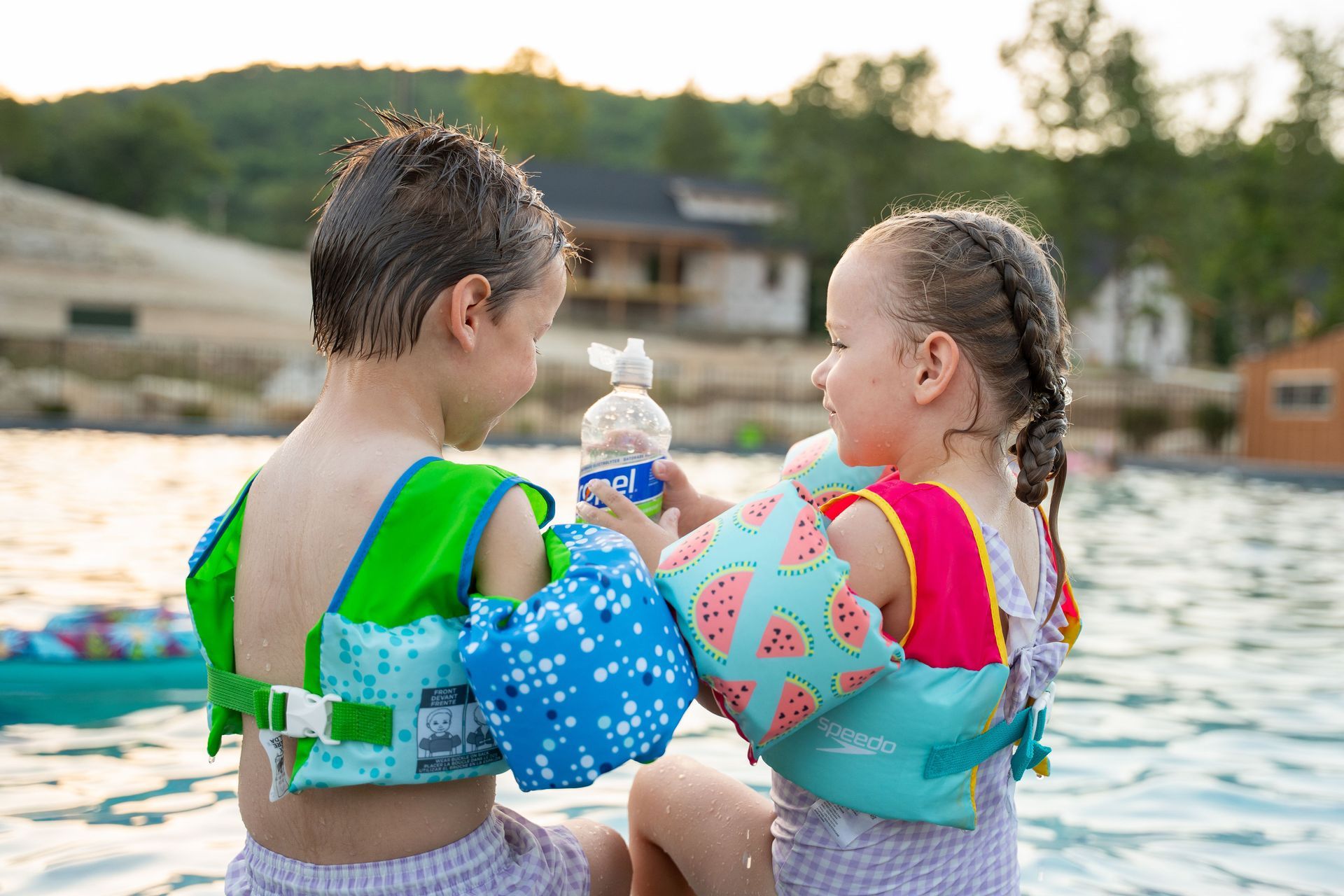 Two children wearing colorful floaties sit at the edge of a lake, facing each other with a water bottle nearby.