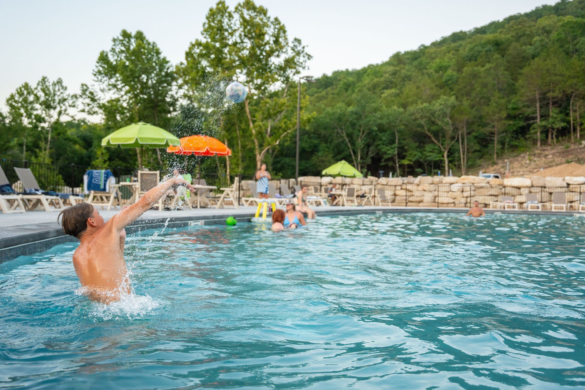 People playing in an outdoor resort pool with a forested hillside in the background