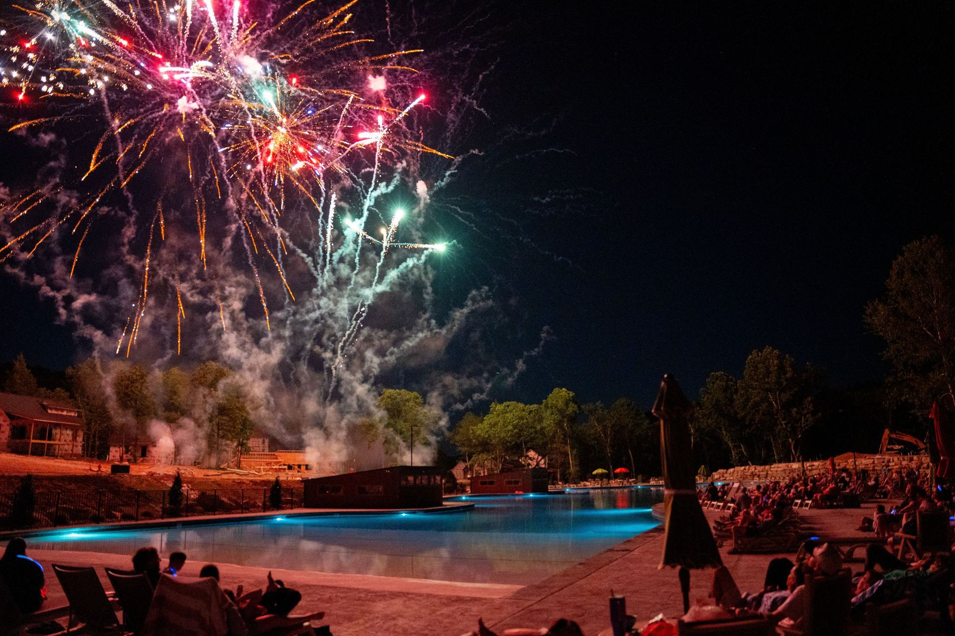 Fireworks bursting over a lit pool at night with a crowd watching from the patio