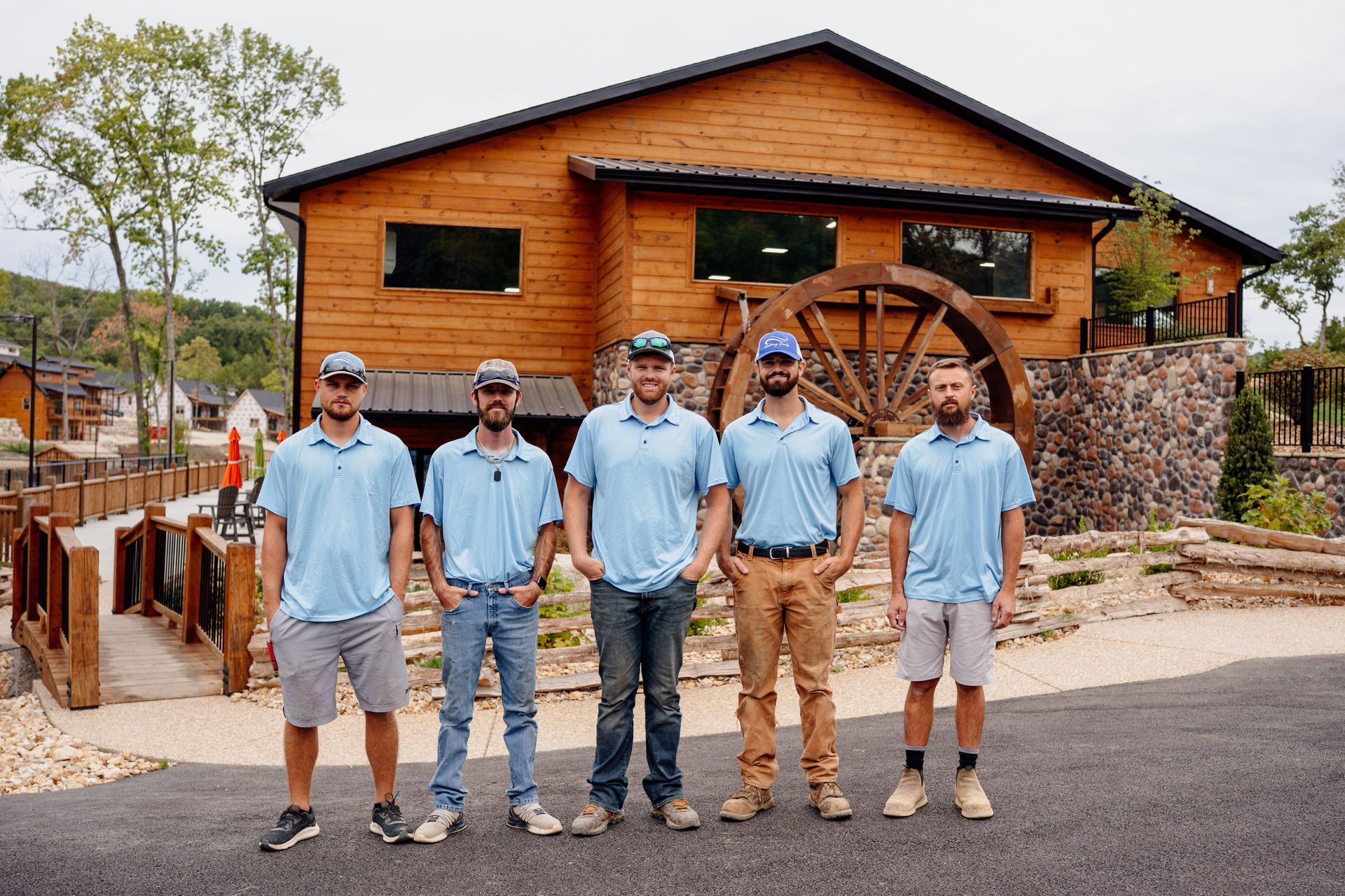 Five men in light blue shirts stand in front of a wooden building on a paved road.