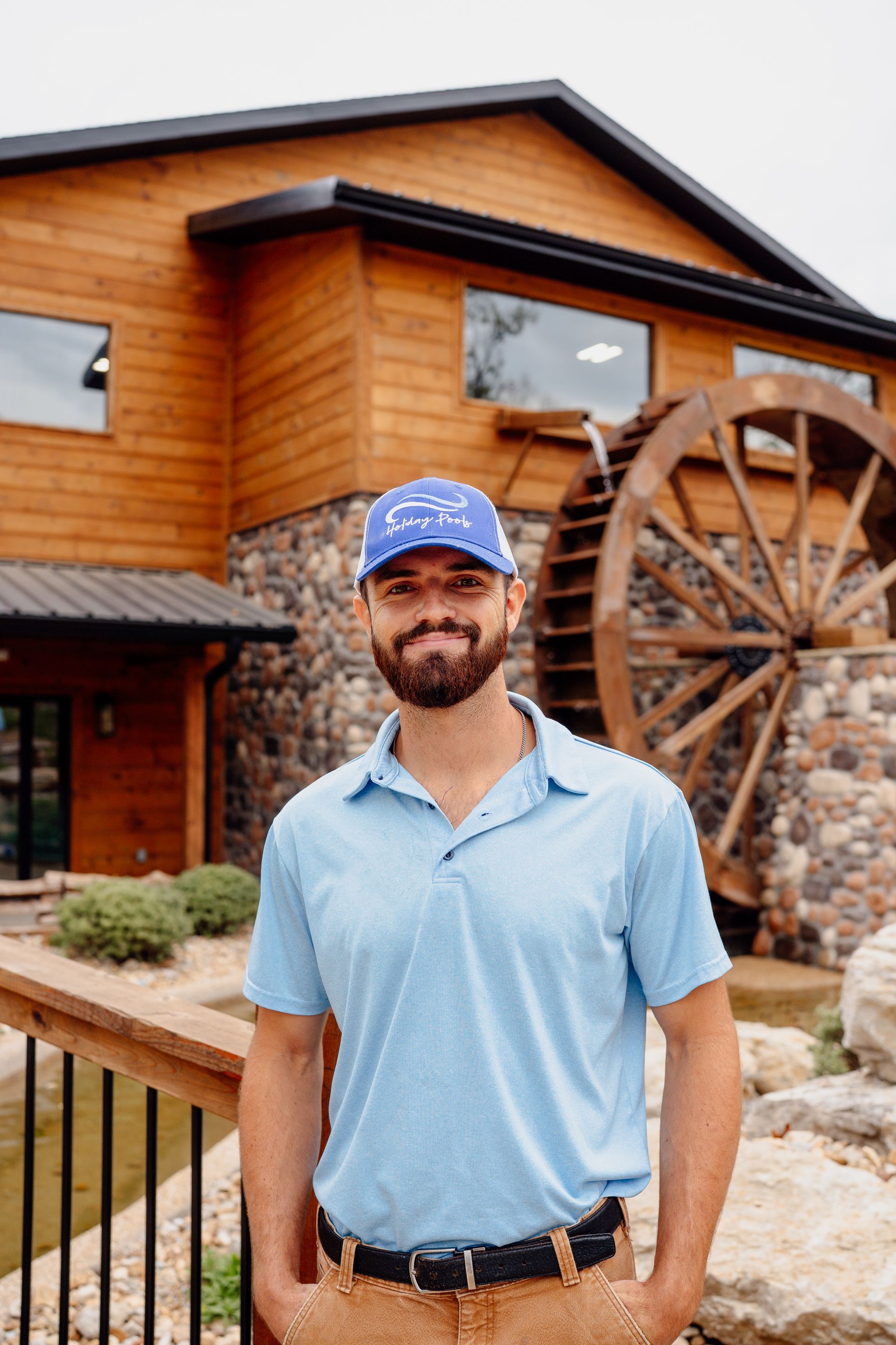 Man in blue shirt and cap standing outside a wooden building with a large water wheel