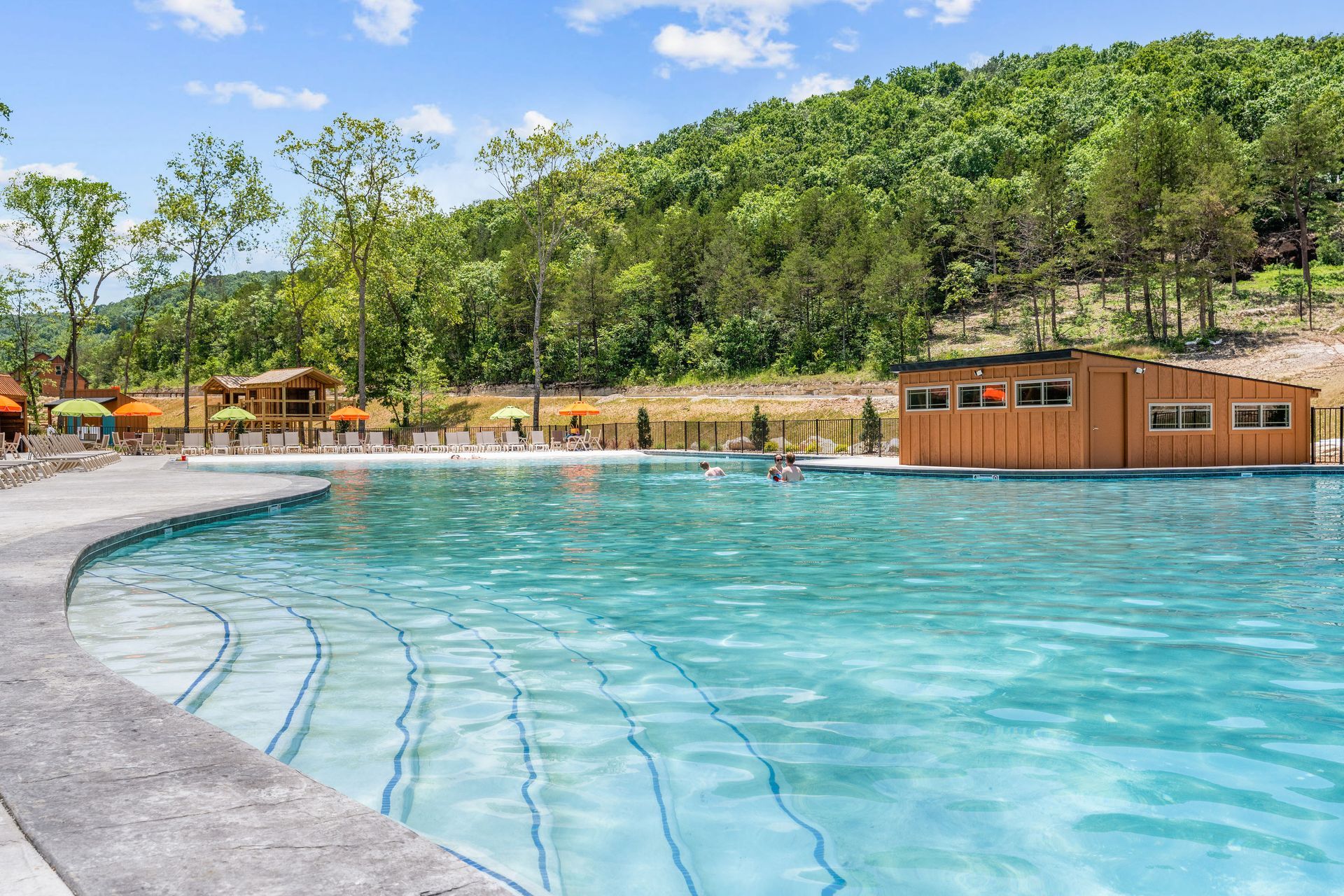 Curved outdoor pool with clear blue water beside a green hillside and wooden cabana on a sunny day
