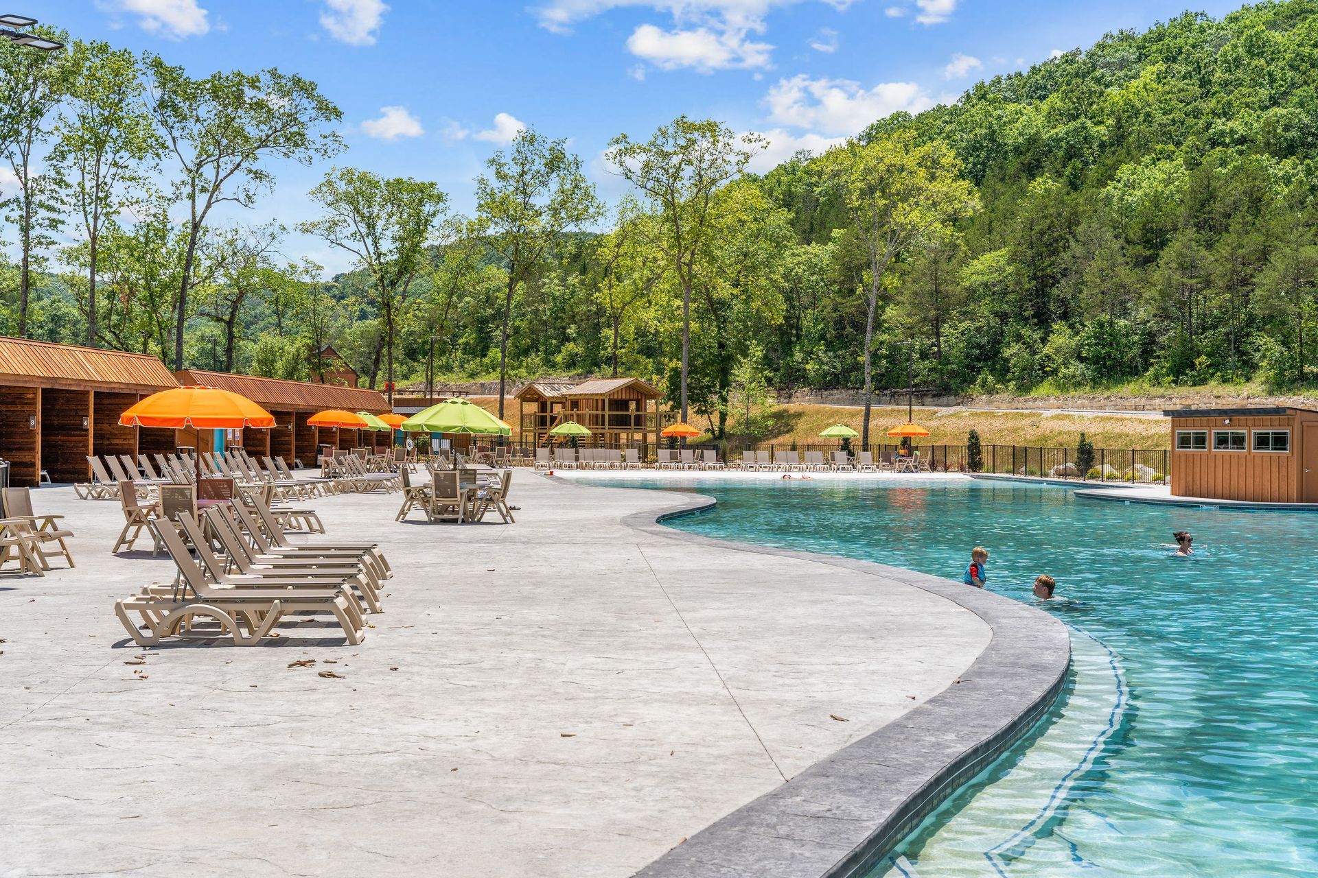Outdoor pool with swimmers, lounge chairs, umbrellas, and trees under a sunny sky