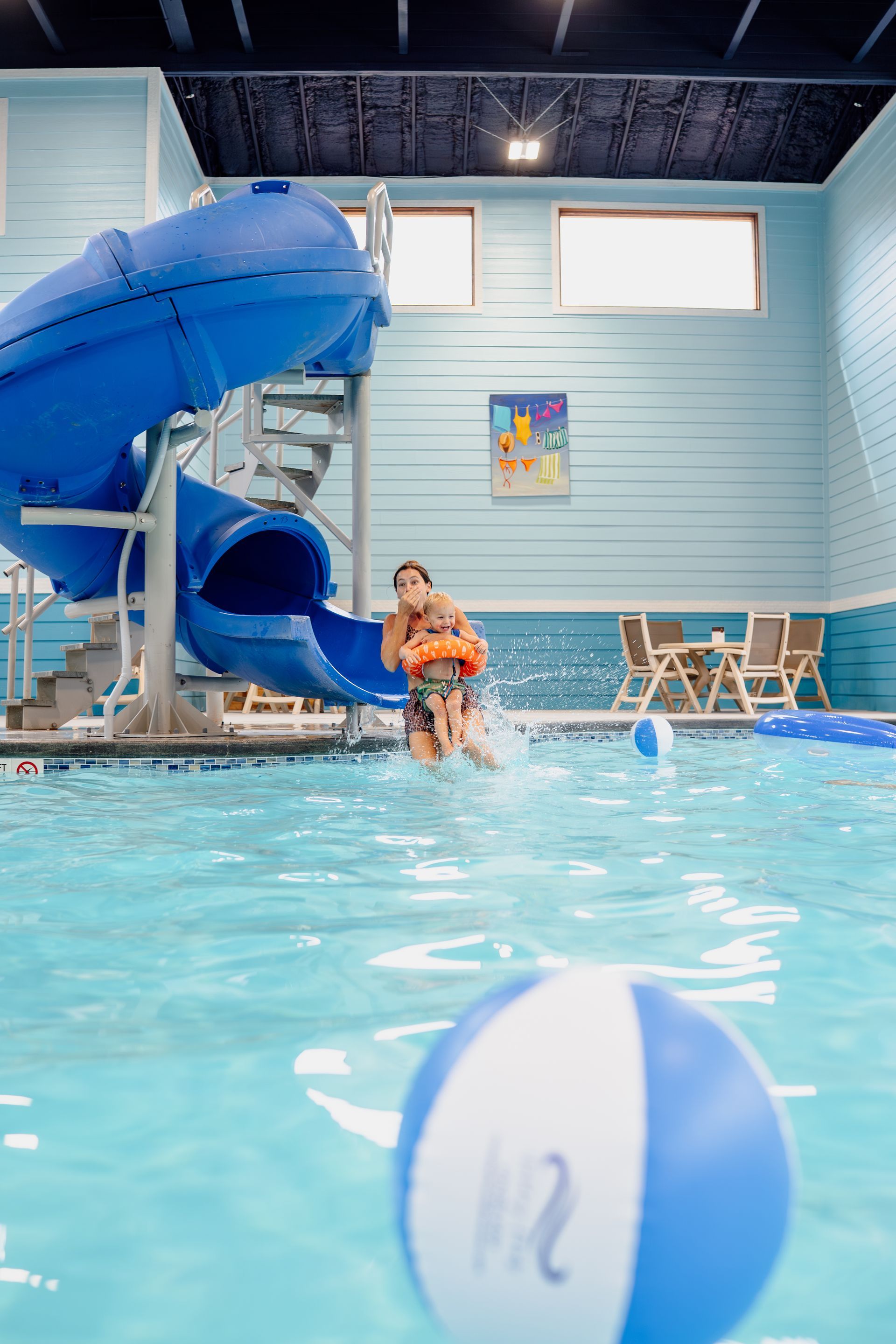 Indoor pool with a blue slide, a person sitting at the edge, and a beach ball in the foreground