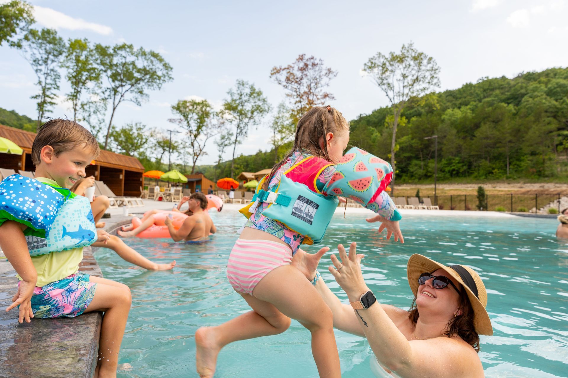People splashing in a pool outdoors, with a child in a pink swimsuit and adults playing in the water.