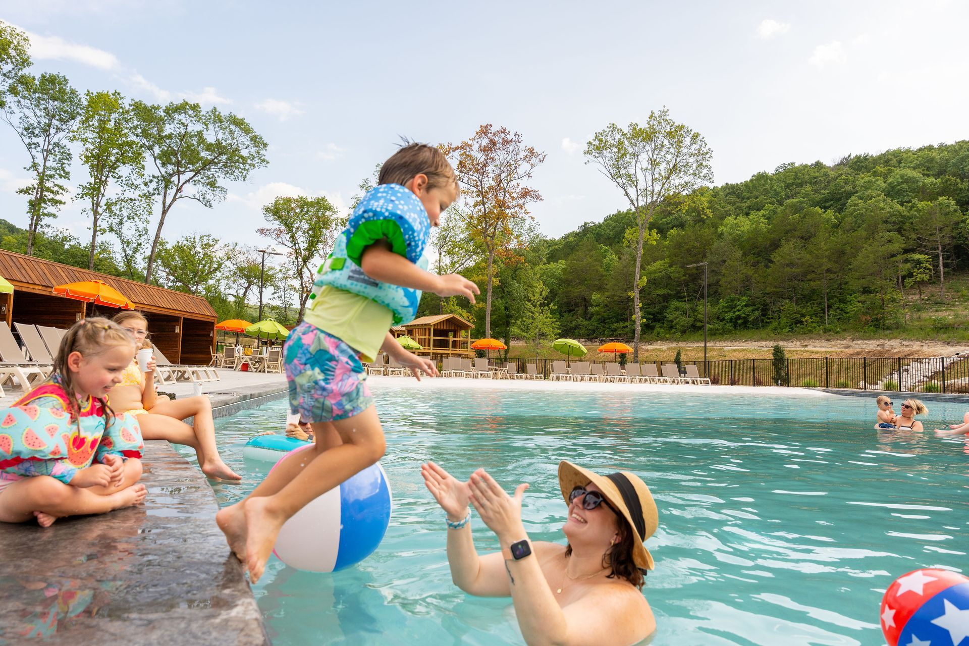 Children playing in a swimming pool with colorful beach balls and a forested hillside nearby