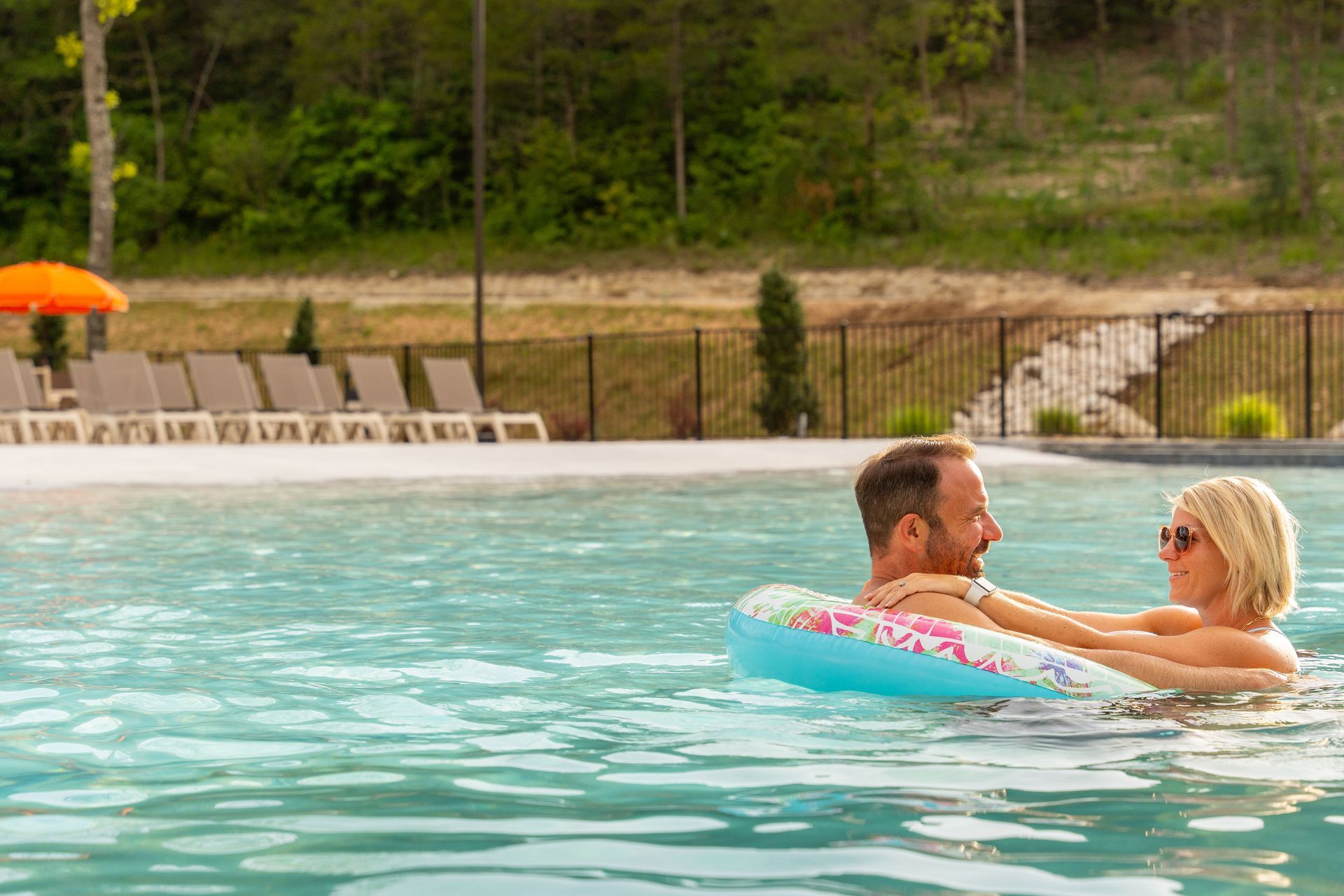 Two people relaxing in a pool with a colorful float, with lounge chairs and trees in the background.