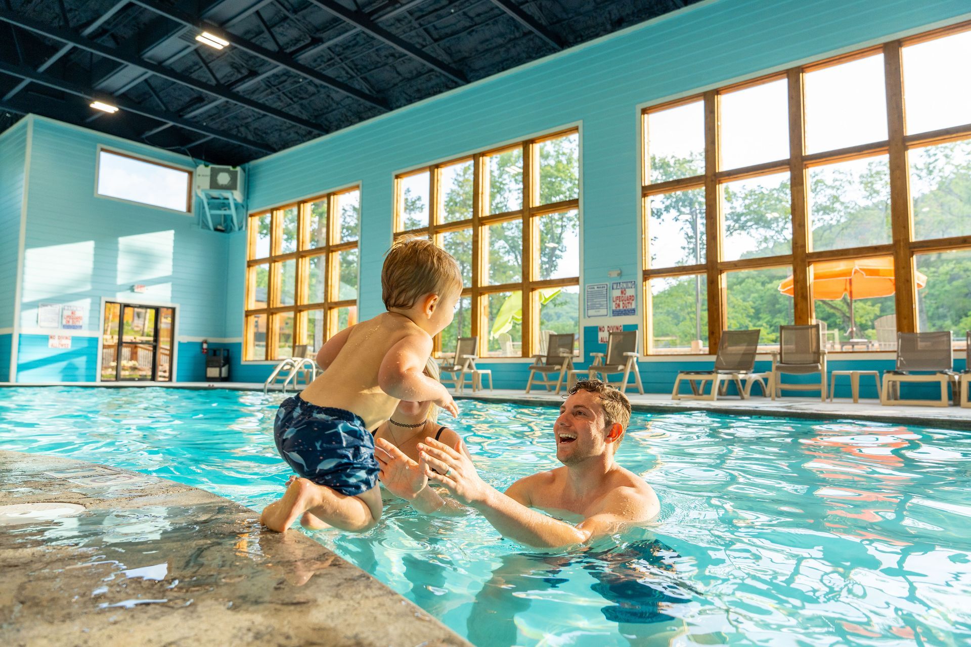 Adult helping child in an indoor pool with bright windows and blue water