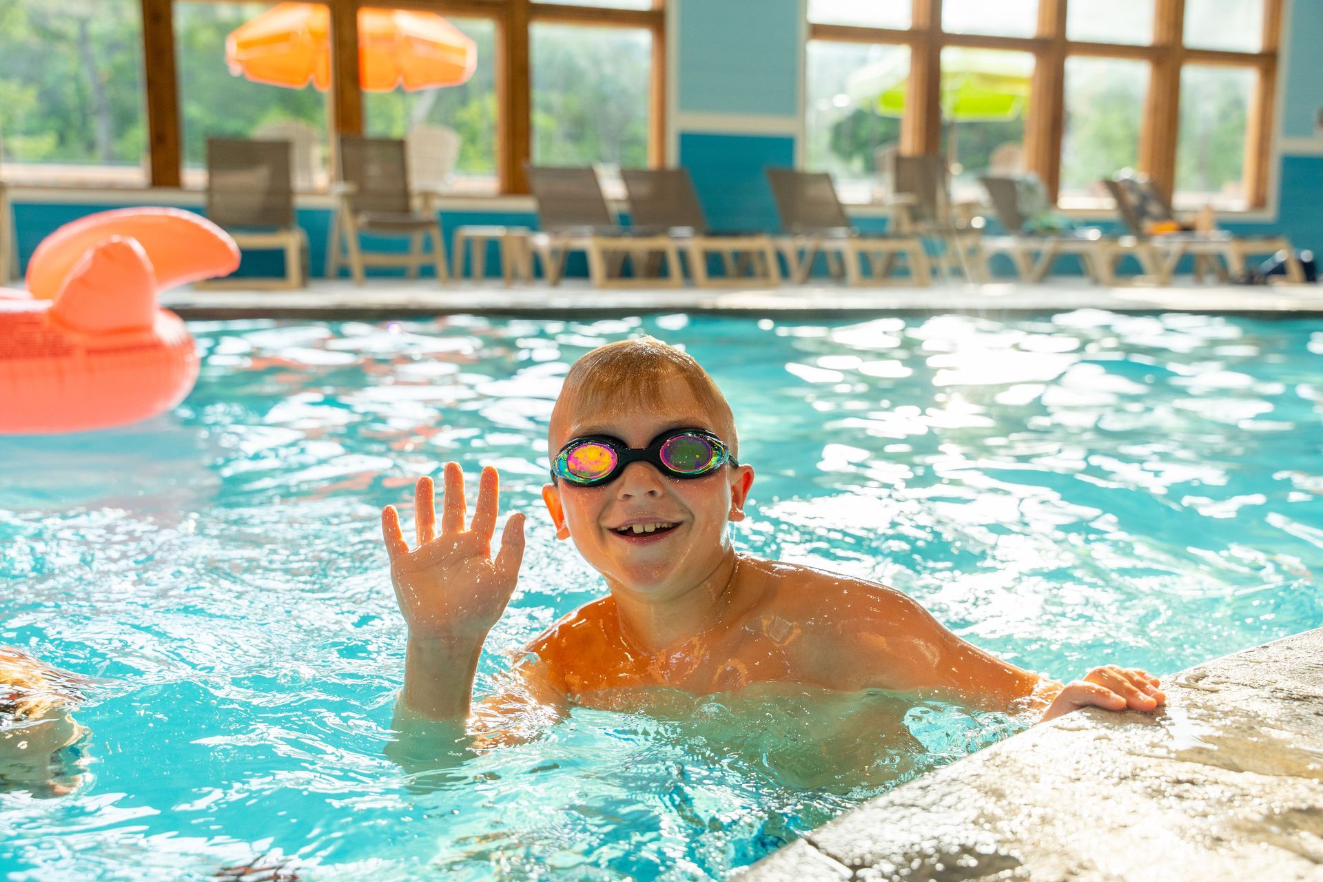 Child in swim goggles waving in an indoor pool, with pool chairs and a large orange float in the background