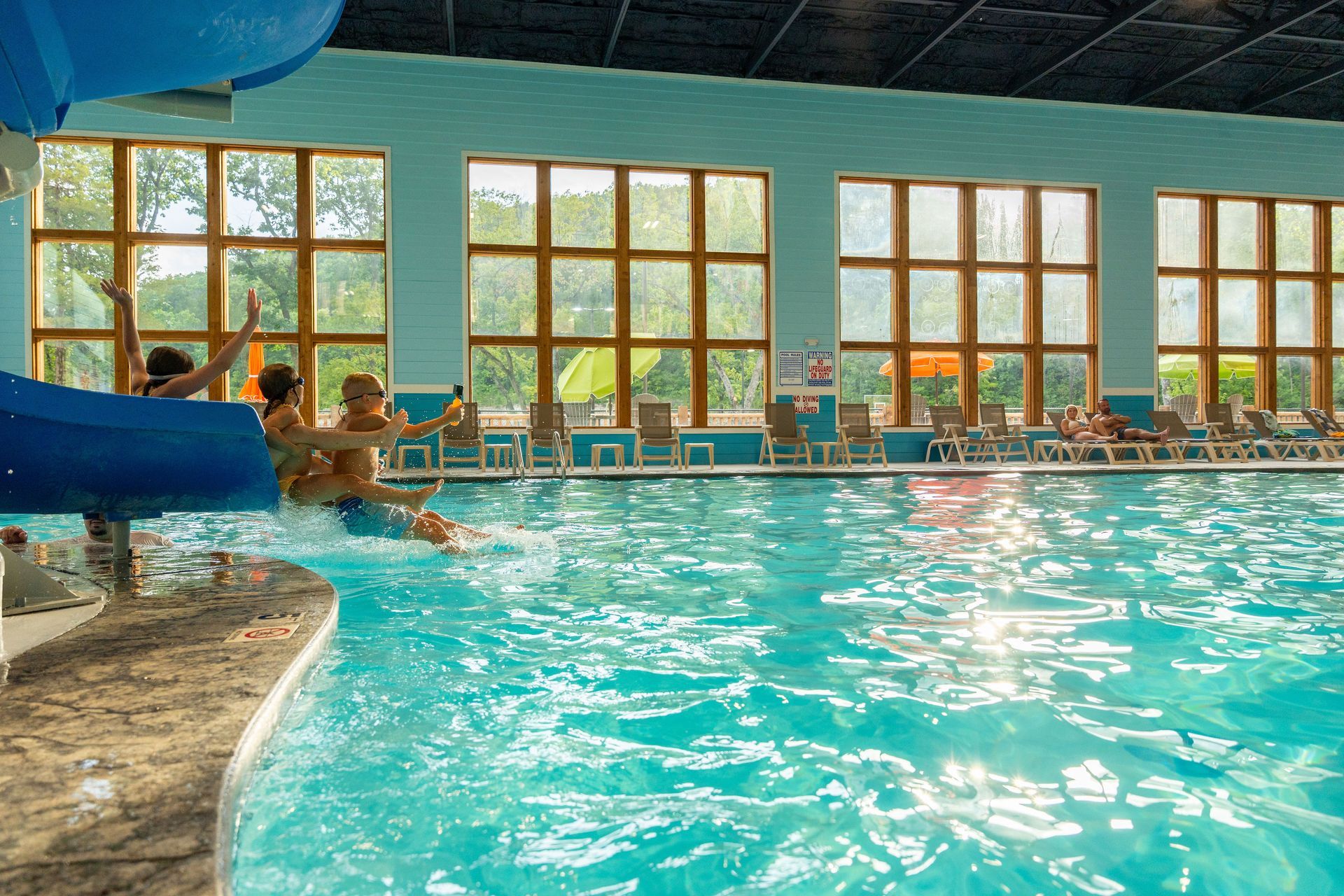 Indoor pool with swimmers near large windows and bright sunlight, turquoise water and a blue slide