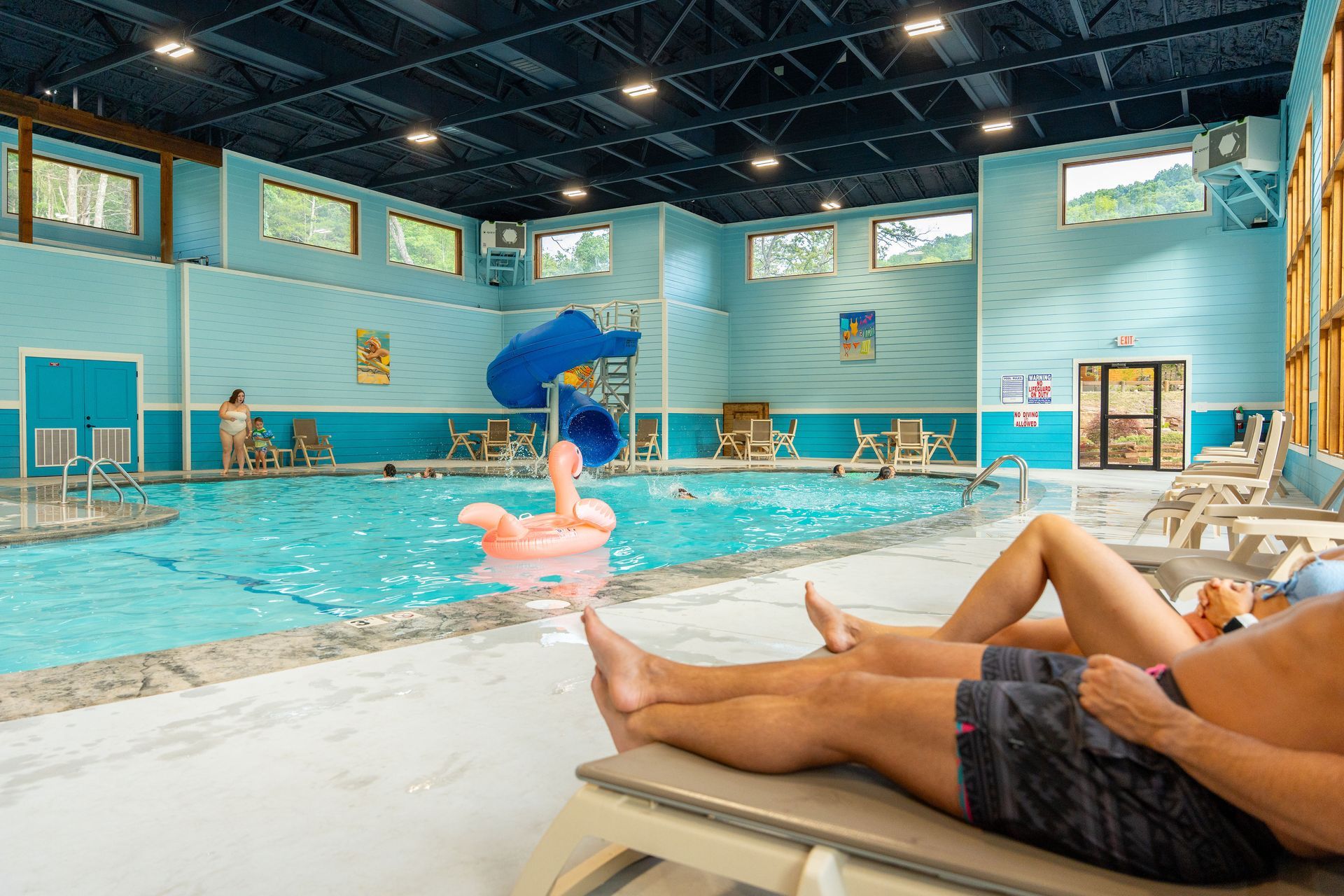 Indoor pool with swimmers, a blue slide, and people lounging on white chairs in the foreground