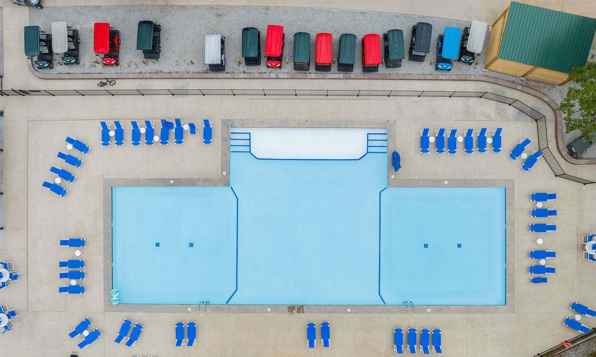 Aerial view of a pool deck with a light blue swimming pool, blue loungers, and parked cars nearby