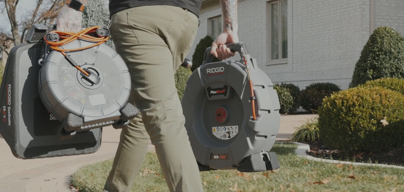 Person carrying two gray coiled-cable containers outdoors on a sunny day.