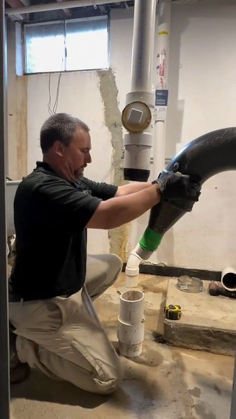 A man installing a curved pipe in a basement. He kneels by a vertical pipe, holding the black pipe with both hands.