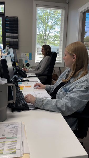 Two women working at desks in an office; one types, the other uses a mouse; both face computer screens.