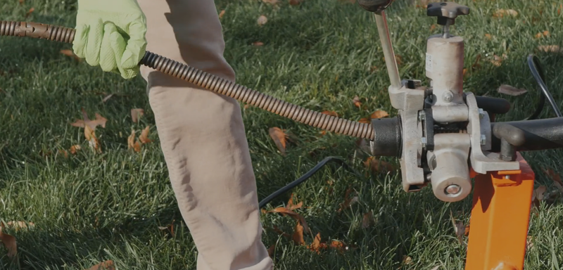 Person using a drain snake on the grass, wearing tan pants and green gloves.
