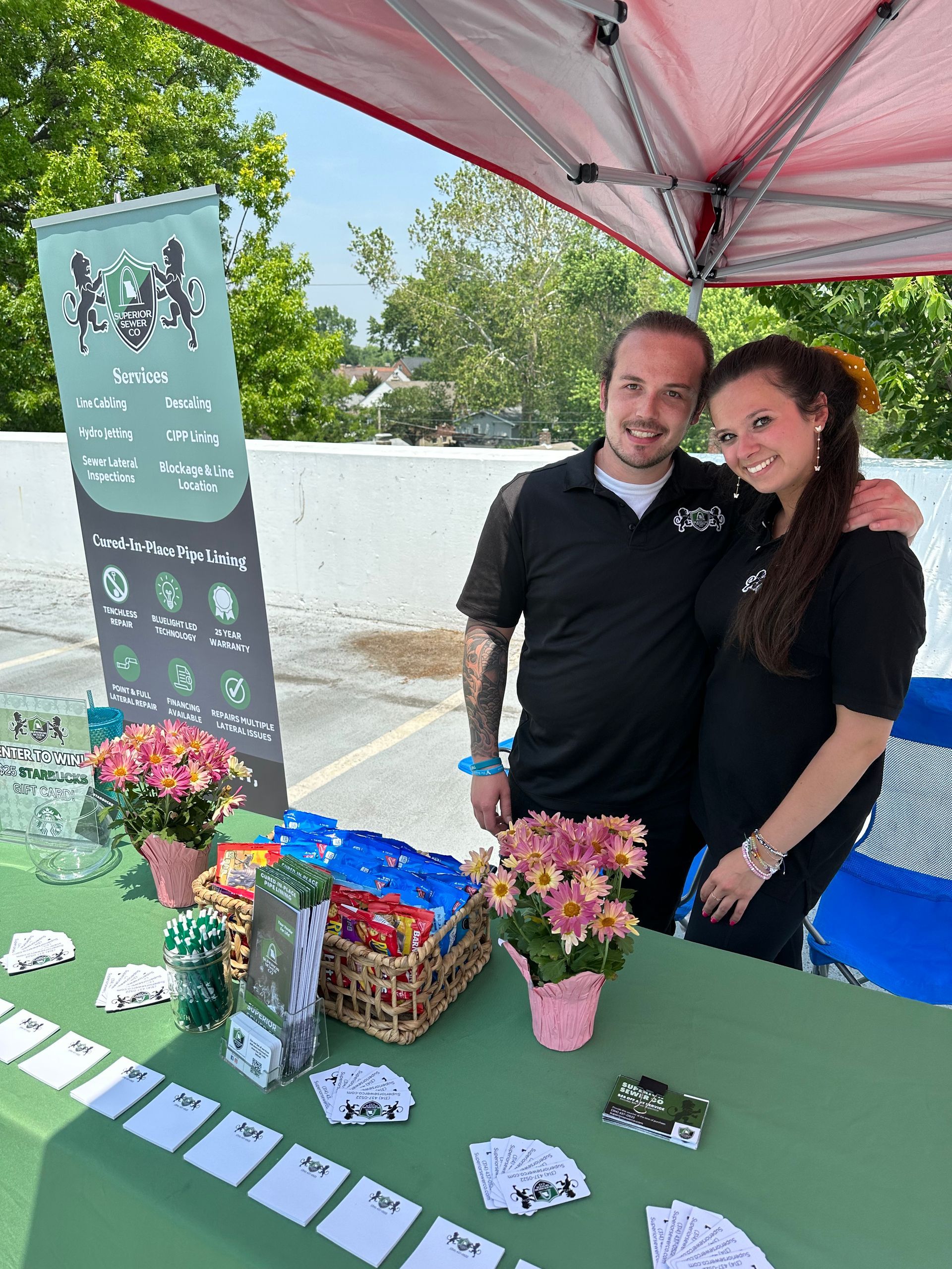 Two people smiling behind a green table with snacks, a sign, and flowers at an outdoor event.