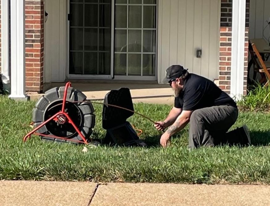 Man kneeling on grass ties a rope to a black speaker near a reel; in front of a building with sliding glass doors.