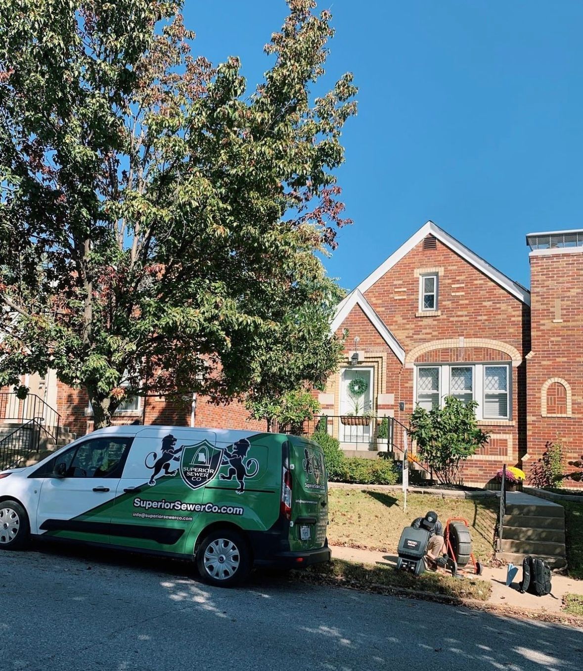 White and green work van parked on a street in front of a brick house on a sunny day.