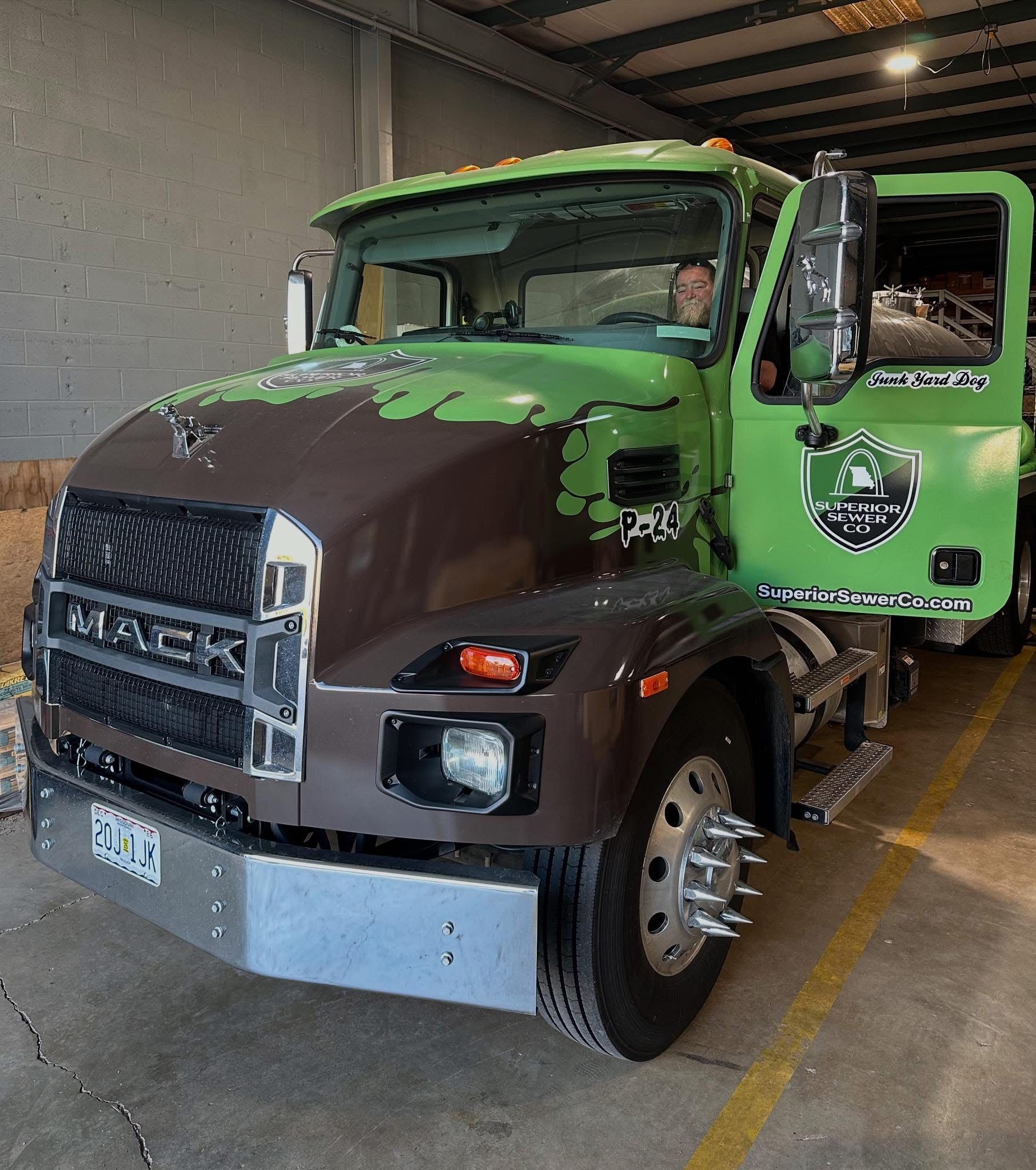Green and brown Mack truck with open door, logo, and driver visible, parked indoors.