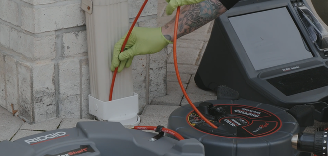 A person in gloves uses a red cable to inspect a downspout near a brick wall, alongside an inspection device.