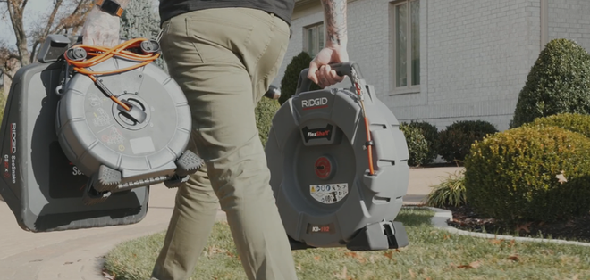 A person carries two gray drain cleaning machines on a lawn in front of a house.