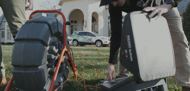 Two people setting up equipment in a yard near a house. One person is operating a machine with a monitor. A car is parked nearby.